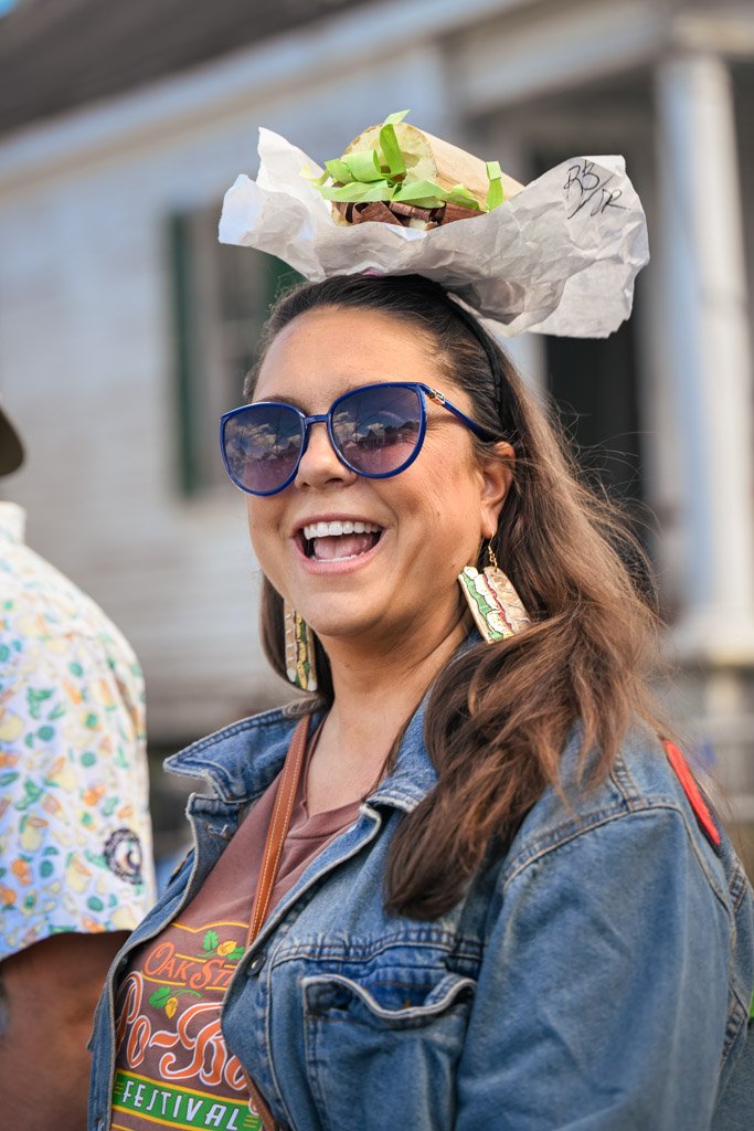A woman wearing large blue sunglasses, earrings, and a denim jacket, laughing, with a sandwich on her head.