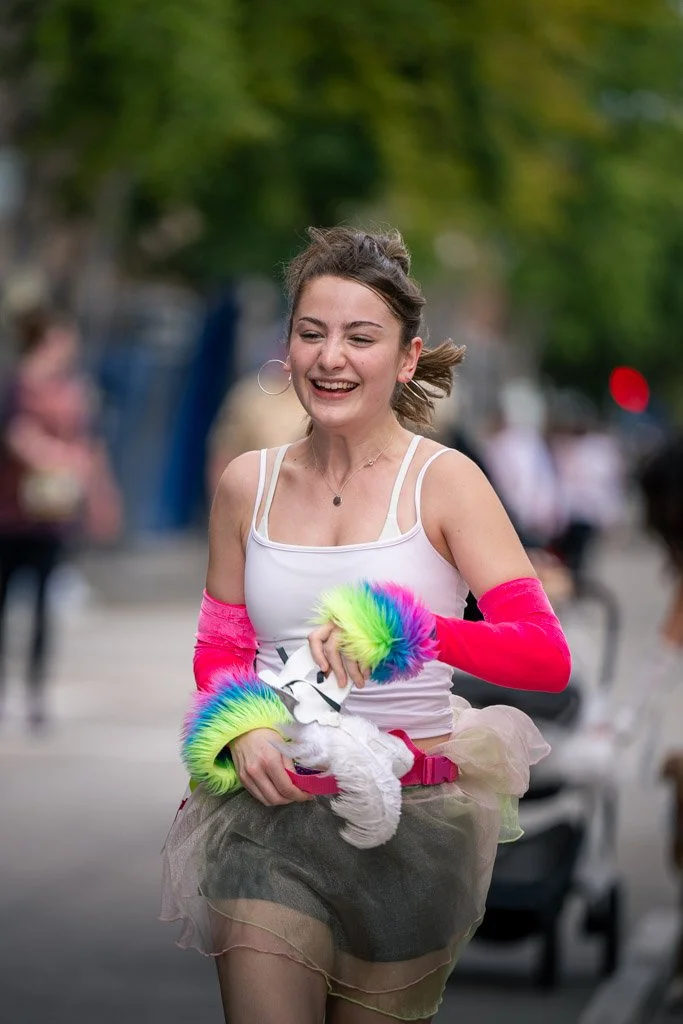 Young woman smiling and running outdoors during a parade or event, wearing colorful furry gloves, a white tank top, a tulle skirt, and holding a small dog.
