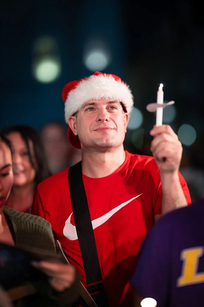 A man wearing a Santa hat and red shirt holding a lit candle in a festive setting with blurred lights in the background.