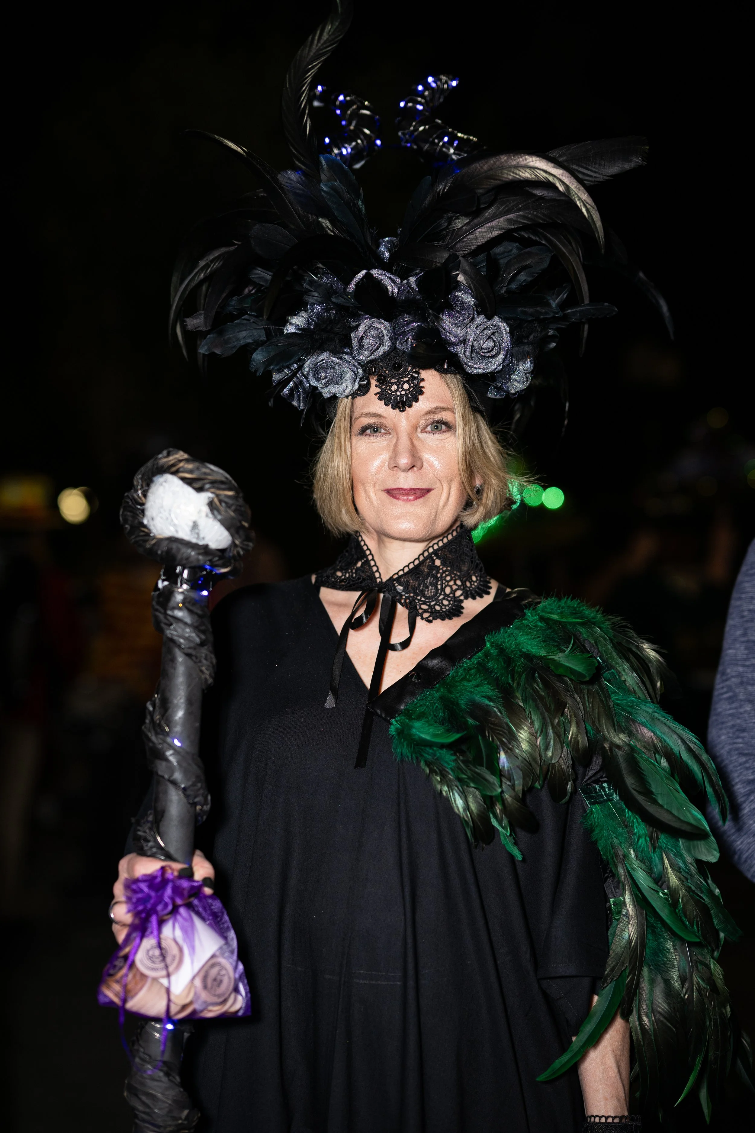 Woman wearing a black dress, a large elaborate headdress with black feathers and flowers, and a feathered shoulder accessory, standing outdoors at night.