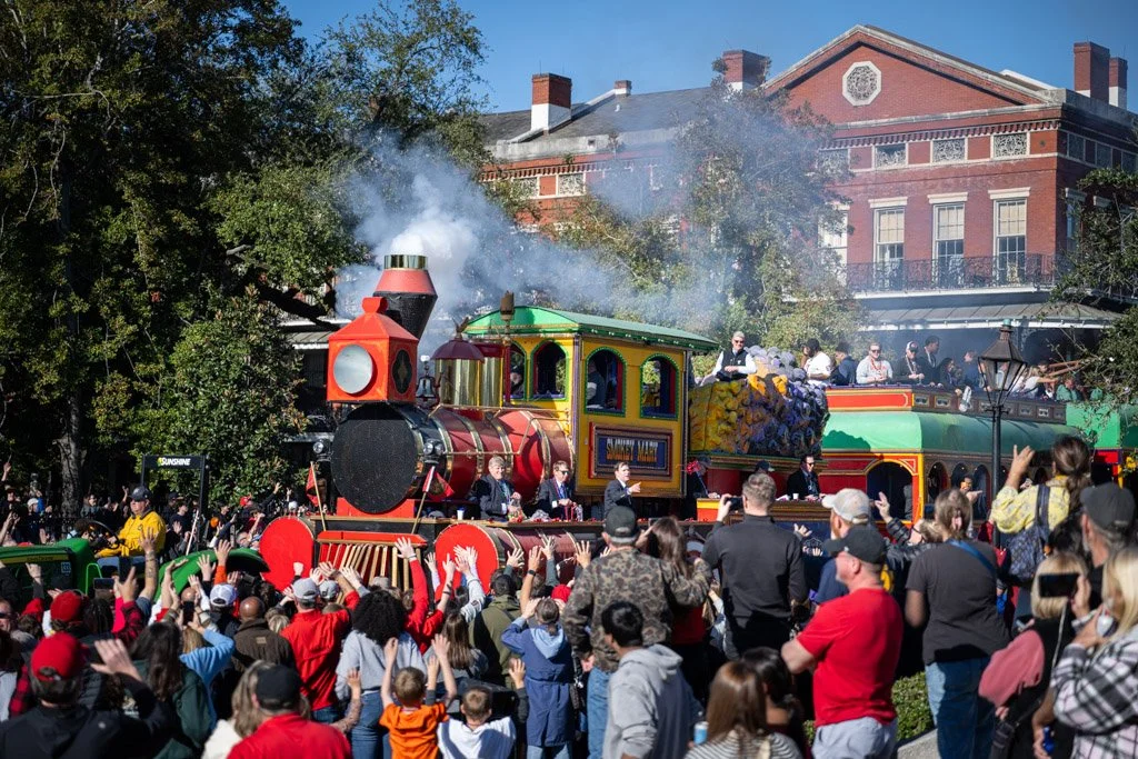 A colorful parade float resembling a train with passengers, surrounded by a crowd of spectators, some waving and taking photos, during a sunny daytime event.