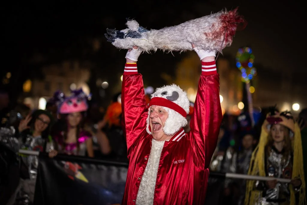 A person dressed in a Santa-themed costume, including a fluffy hat with a koala face design, holding a plush chicken above their head during a nighttime parade or festival, with many people in costumes and holiday lights in the background.
