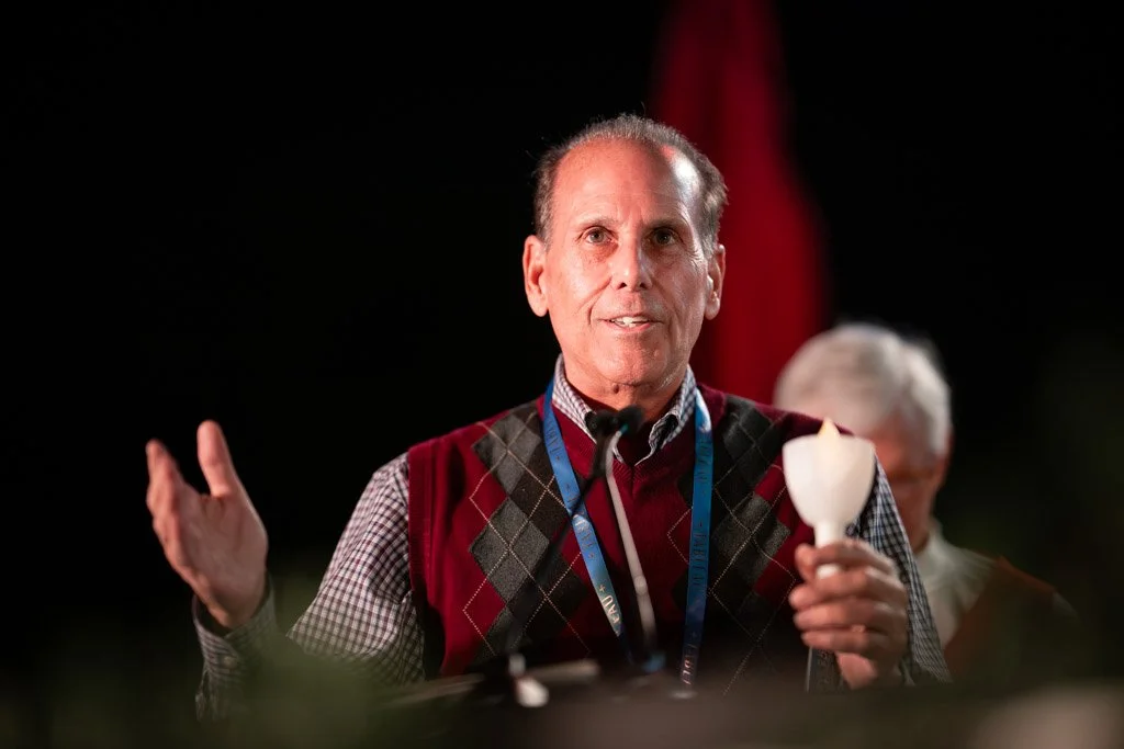A man in a red and gray argyle vest speaking into a microphone at an event, with another person partially visible in the background.