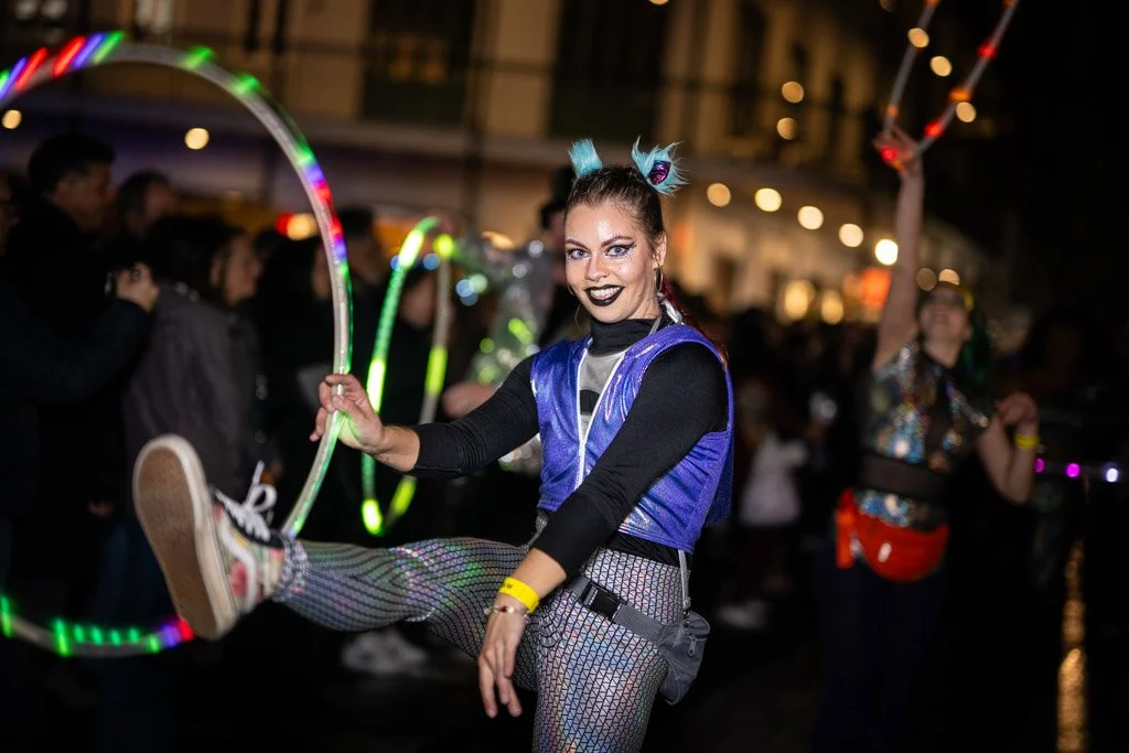 A woman dressed in colorful festival attire, with her hair styled in pigtails and makeup, is dancing with LED hula hoops at a nighttime outdoor event surrounded by a crowd.