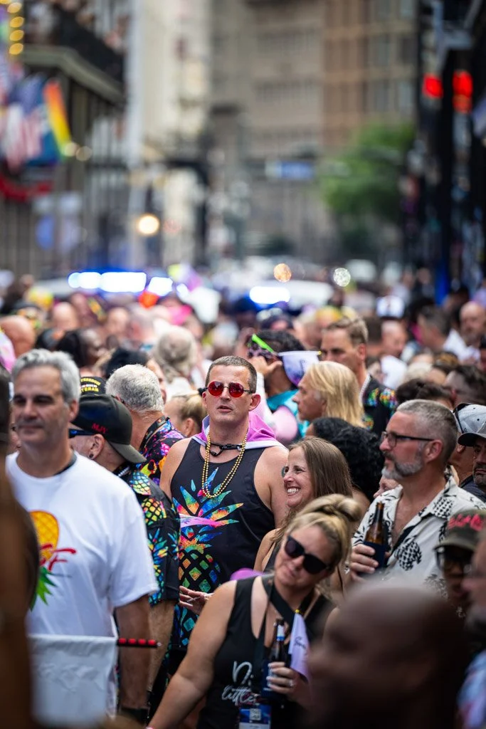 Crowd of people at an outdoor event, some wearing colorful clothing, with buildings and city street in the background.