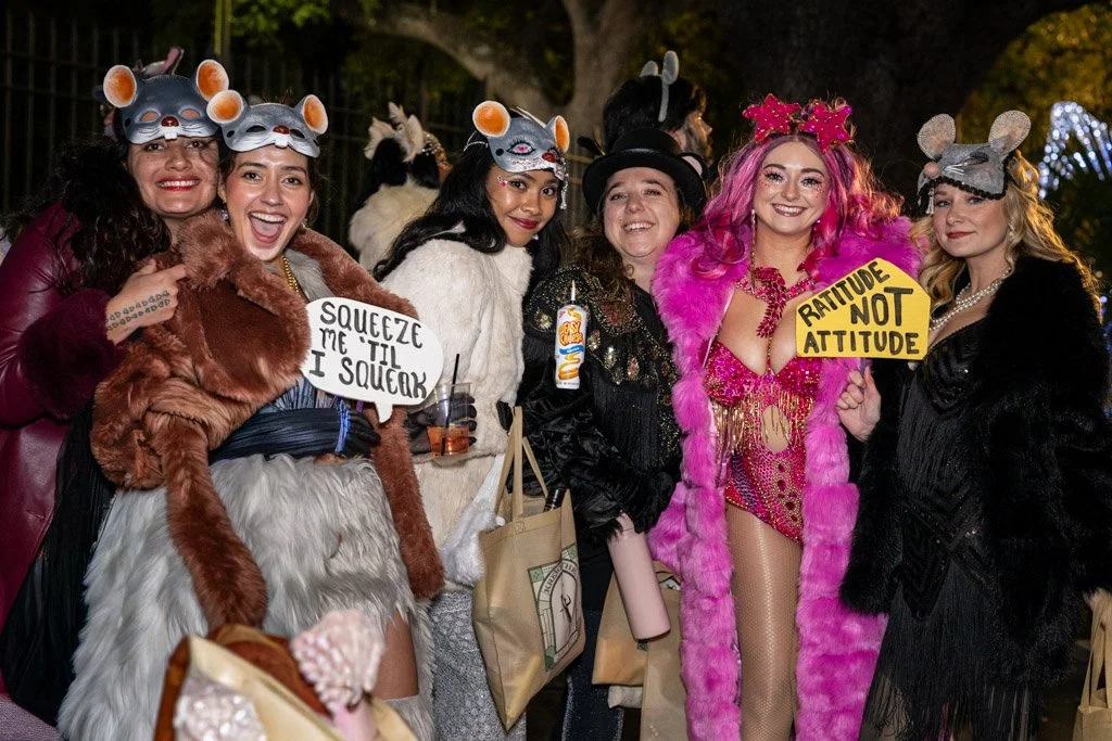 Group of women dressed in whimsical costumes, some wearing mouse masks and others with colorful accessories, smiling and holding signs at a festive outdoor event at night.