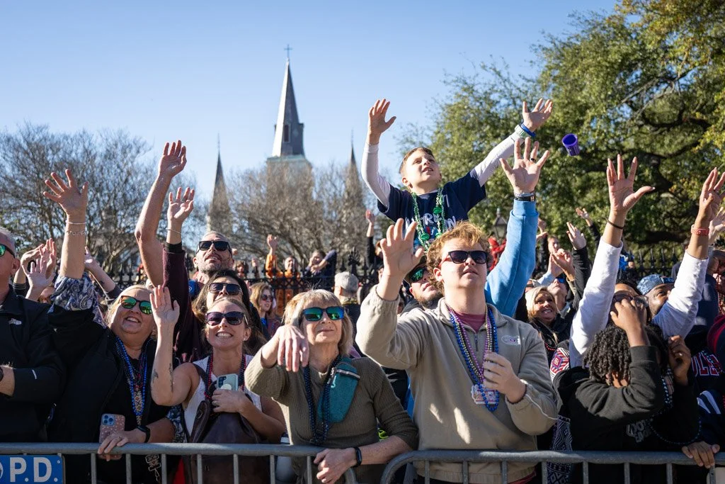 Crowd of people celebrating outdoors, some wearing sunglasses and beads, raising their hands with a church steeple and trees in the background.