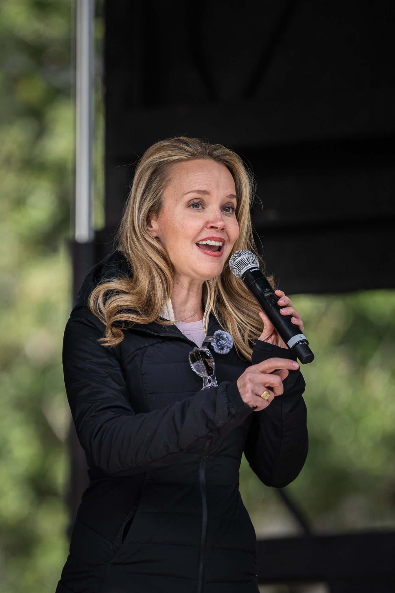 A woman with blonde hair speaking into a microphone at an outdoor event, wearing a black jacket and sunglasses hanging from her jacket.