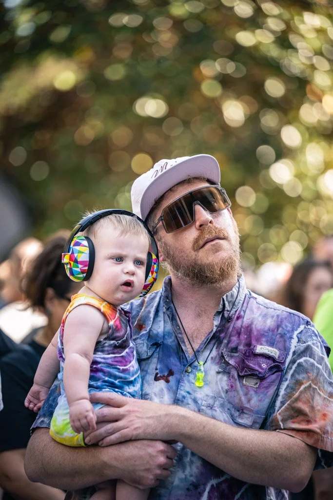 A man with a beard, wearing sunglasses, a cap, and a tie-dye shirt, holds a young child wearing large headphones and a colorful tie-dye outfit, outdoors with a blurred background of trees and people.