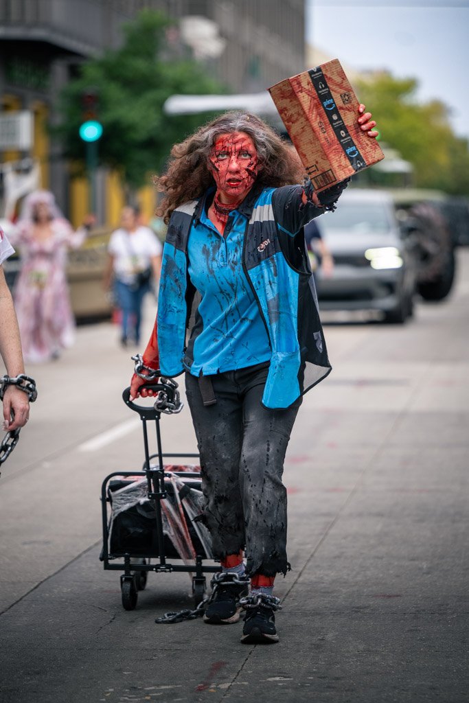 A woman dressed as a zombie with a gory face makeup, wearing a blue jacket, walking on a city street pulling a shopping cart, holding a cardboard box in one hand.