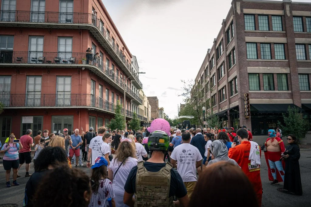Crowd gathered on a city street during daytime, with multi-story buildings on either side, some people on balconies, and individuals in costumes among the crowd.