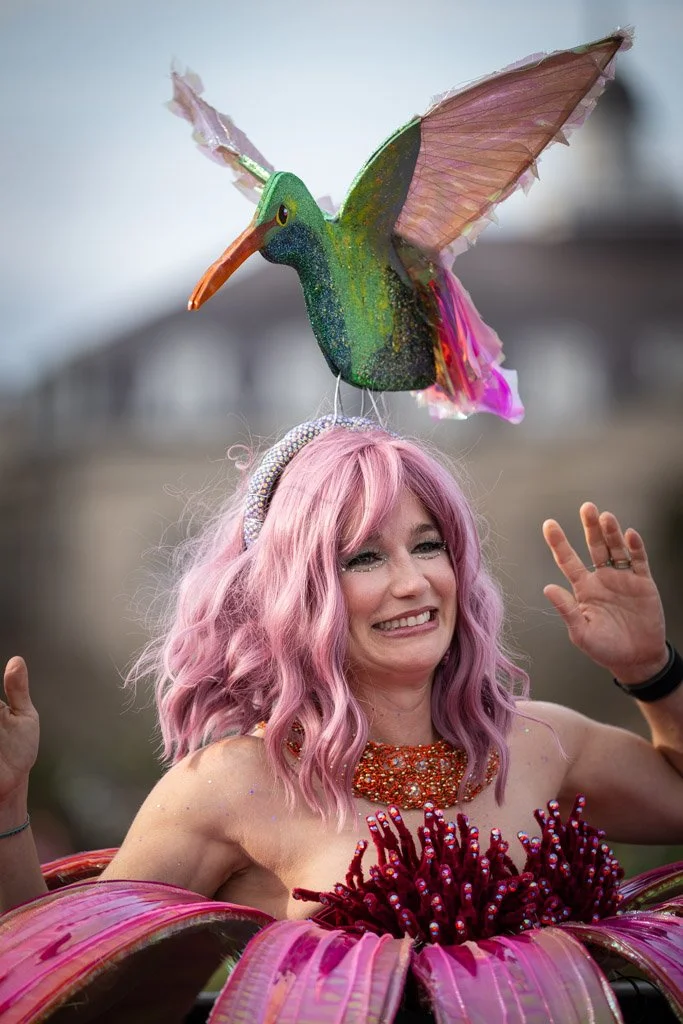 A woman with pink hair wearing a vibrant costume with a large flower-like collar, smiling and raising her hands, with a colorful hummingbird-shaped hat on her head.