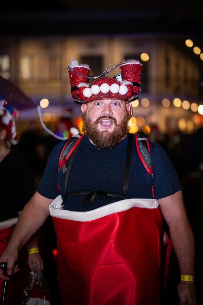 Person wearing a Christmas-themed hat with red glitter, white pom-poms, and white balls, at a festive event at night.