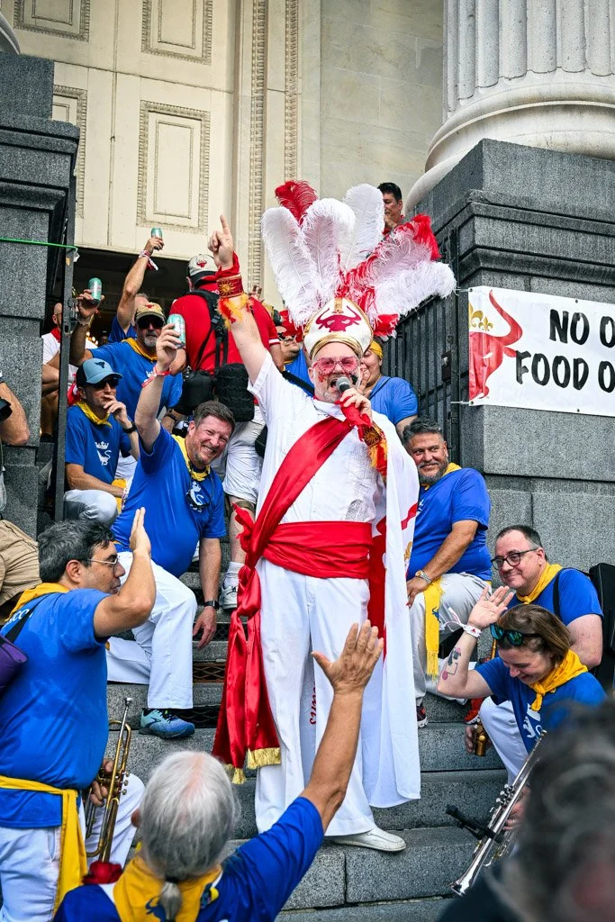 A person dressed as a Roman or Greek deity, wearing a white toga with red sash and a tall feathered headdress, speaking into a microphone on steps surrounded by a group of people in blue shirts celebrating at an outdoor event.