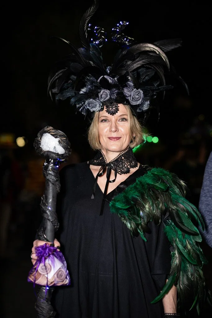 A woman dressed in a black costume with feathered accessories, wearing an elaborate headpiece decorated with black feathers, roses, and blue lights, holding a staff with a purple and white cloth.