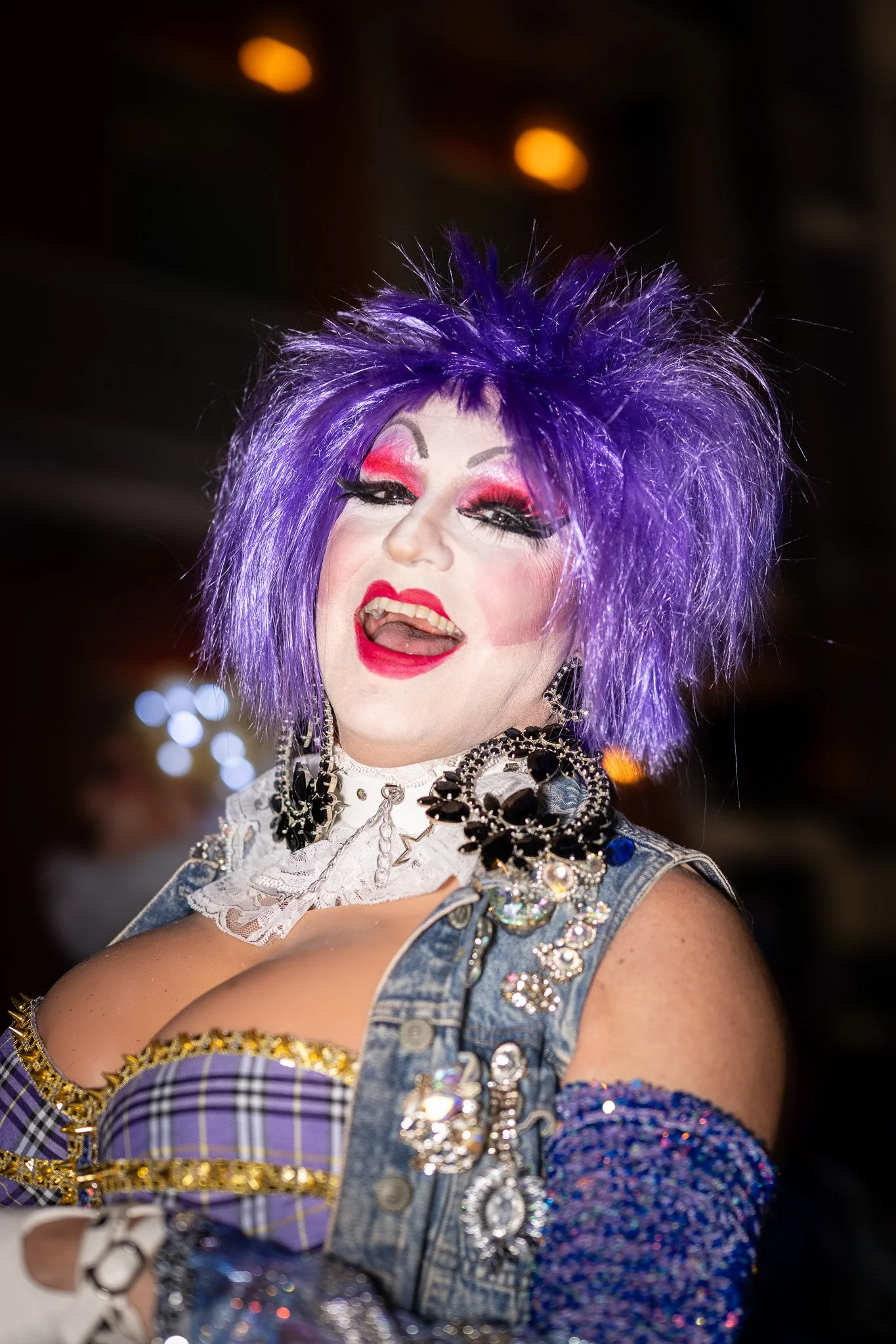A person with purple spiky wig and vibrant makeup smiling and posing with their tongue slightly out, dressed in a colorful, embellished outfit.