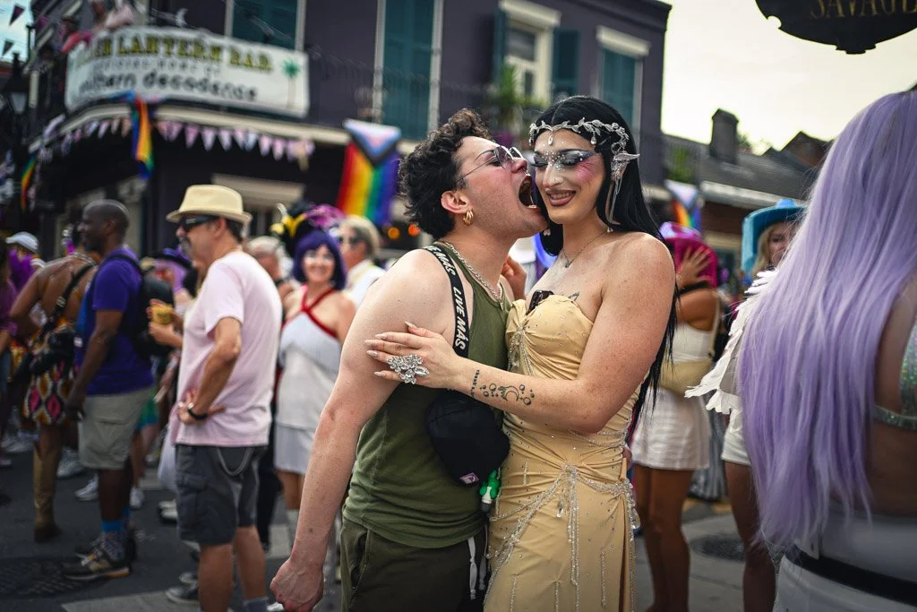 Two people celebrating at a pride parade, one wearing green and the other in a yellow dress with glitter makeup and accessories, surrounded by a crowd of diverse parade attendees with rainbow decorations.