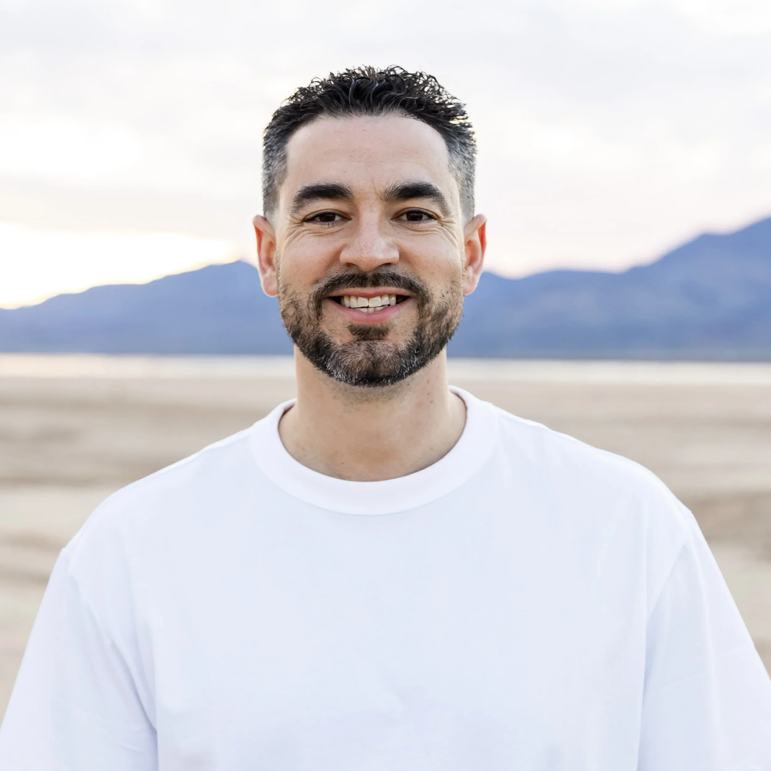 A smiling man with dark hair and a beard outdoors, with mountains and a body of water in the background, wearing a white t-shirt.
