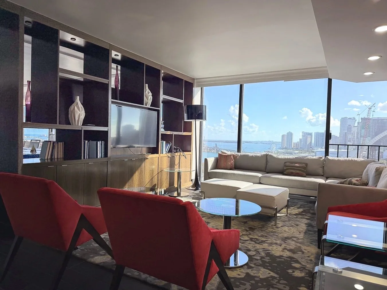 Living room with large windows showing city and water view, beige sectional sofa, red chairs, and dark wood shelving unit with a TV.
