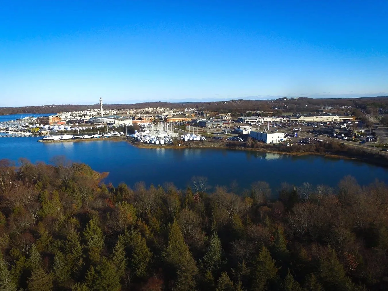 Aerial view of a marina with boats covered in white tarps, next to a body of water, with a parking lot and commercial buildings in the background, and wooded area in the foreground.