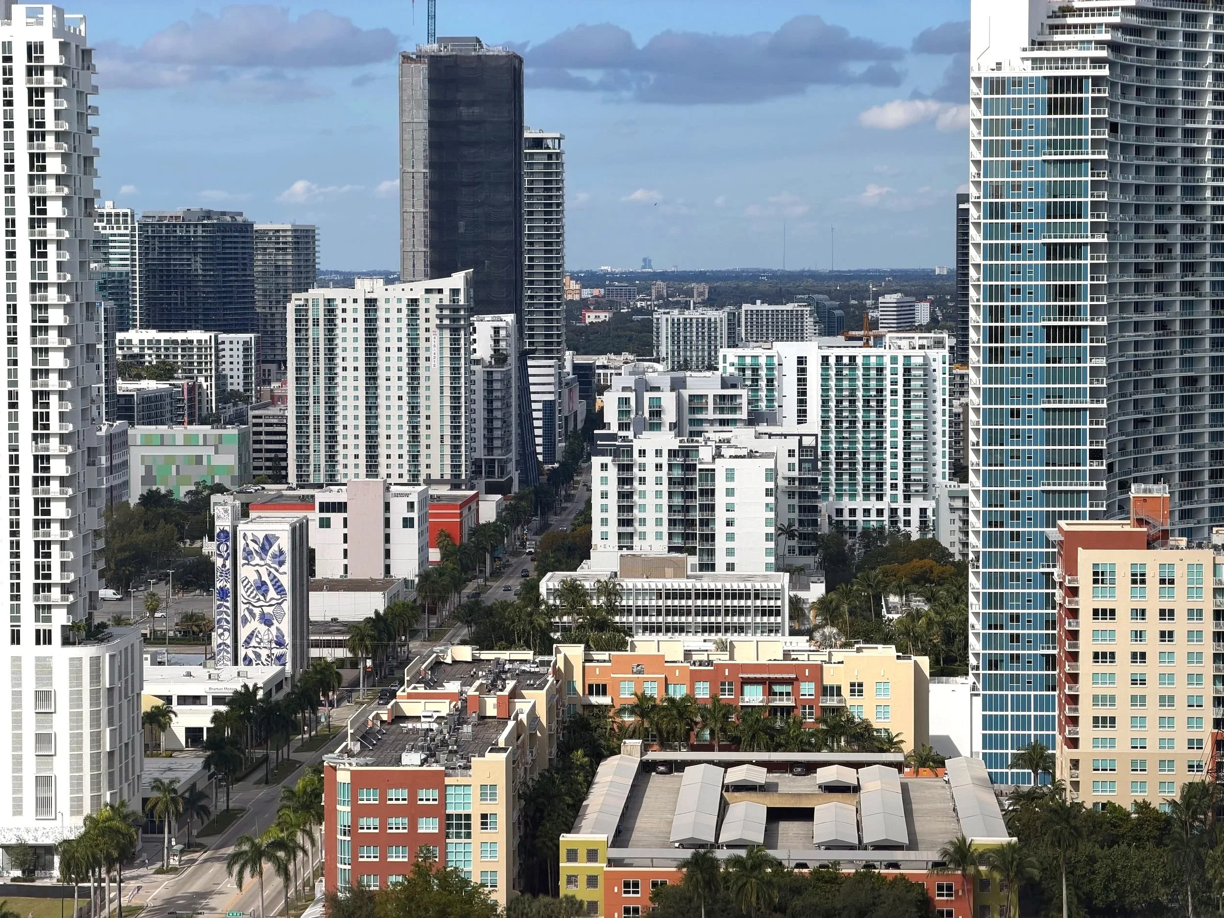 A cityscape with numerous high-rise buildings, mostly white or light-colored, with a tall dark building under construction in the center. There are palm trees lining the streets and a partly cloudy sky overhead.