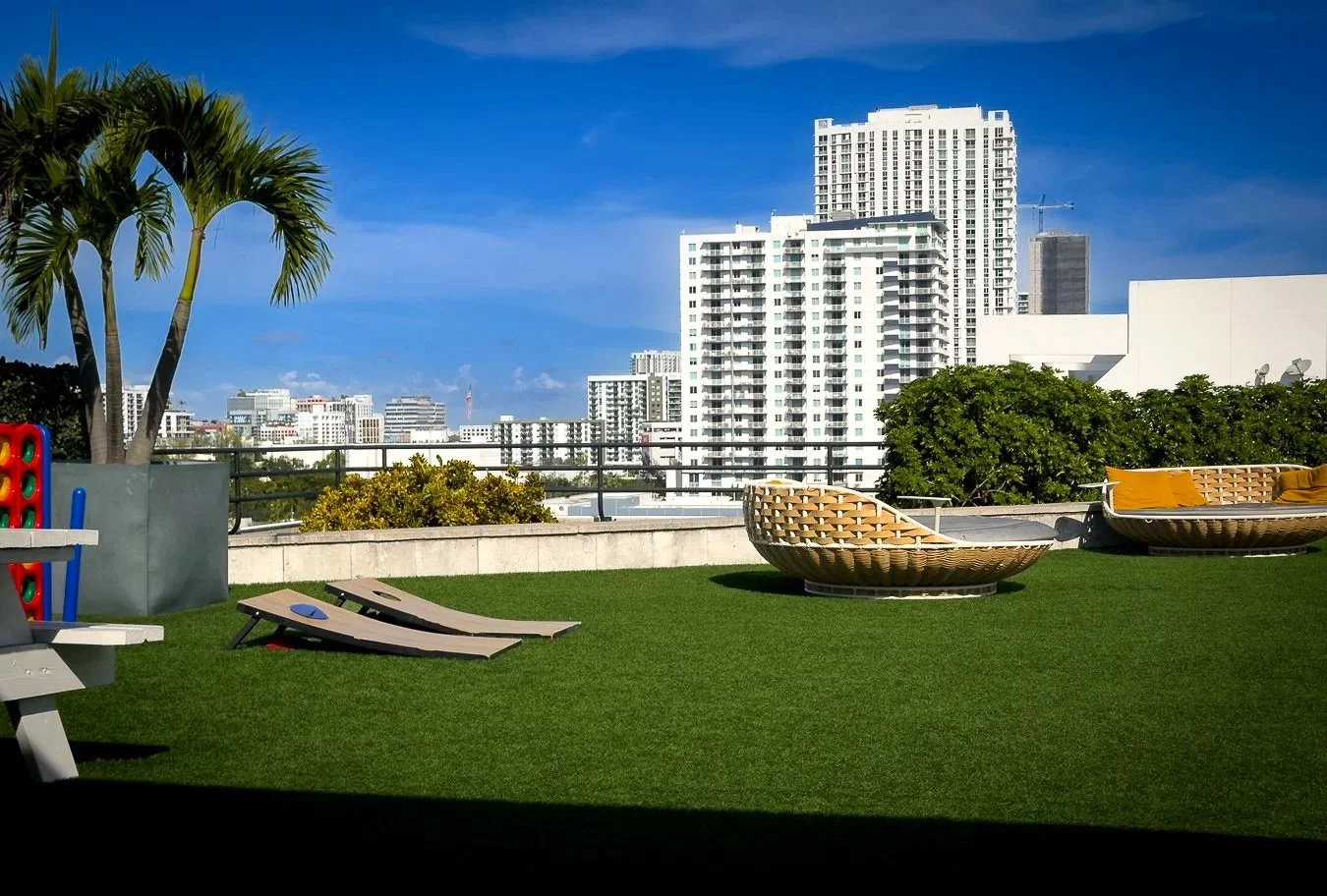 Rooftop garden with lounge chairs, outdoor seating, a palm tree, green bushes, and city buildings in the background during the daytime.
