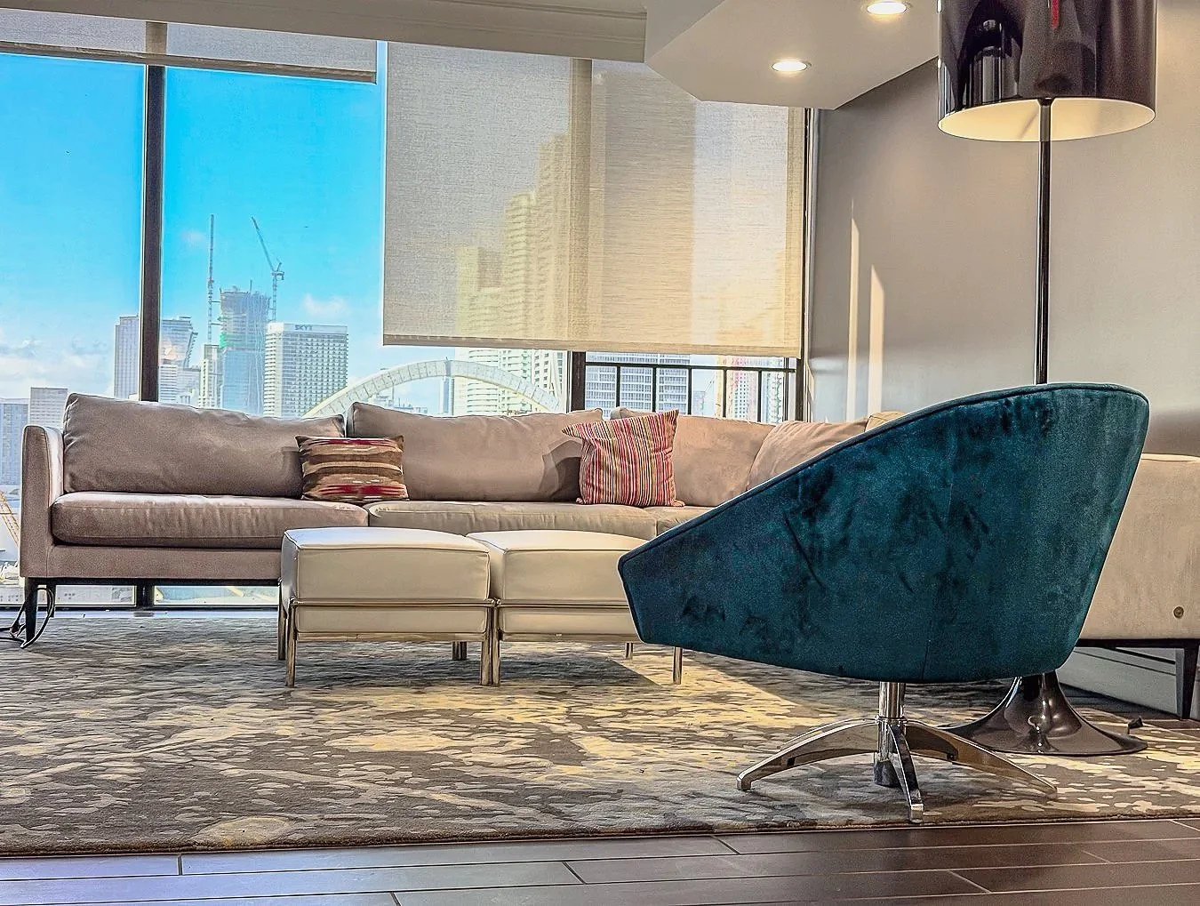 Modern living room with large window showing city skyline, beige sofa with cushions, white ottoman, teal armchair, and a rug.