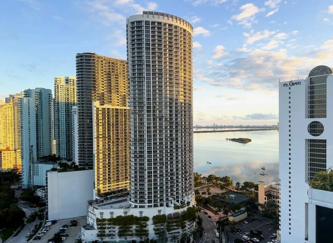 Skyline of tall modern high-rise buildings along a river with a small island, palm trees, and a parking lot in the foreground, under a partly cloudy sky during sunset.