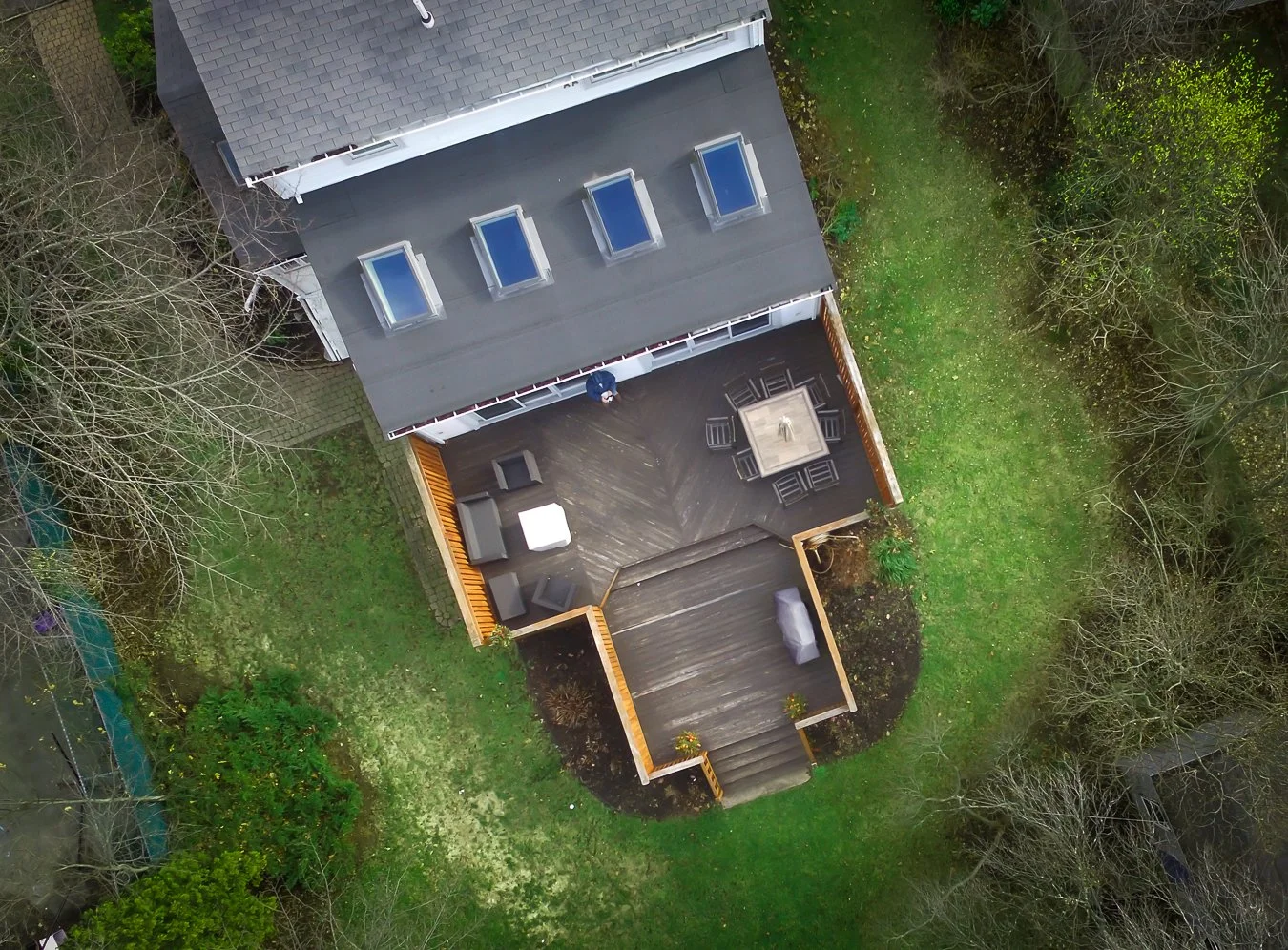 An aerial view of a backyard deck attached to a house, with outdoor furniture including chairs, table, and a lounging area, surrounded by grass, trees, and hedges.