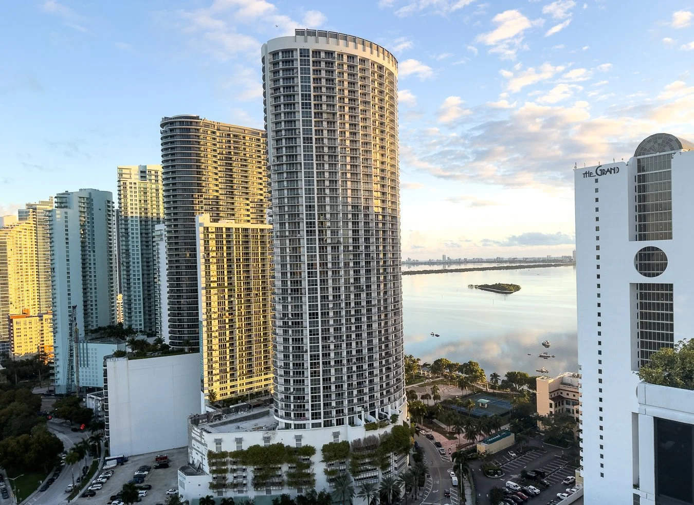 Skyline of tall modern high-rise buildings along a river with a small island, palm trees, and a parking lot in the foreground, under a partly cloudy sky during sunset.