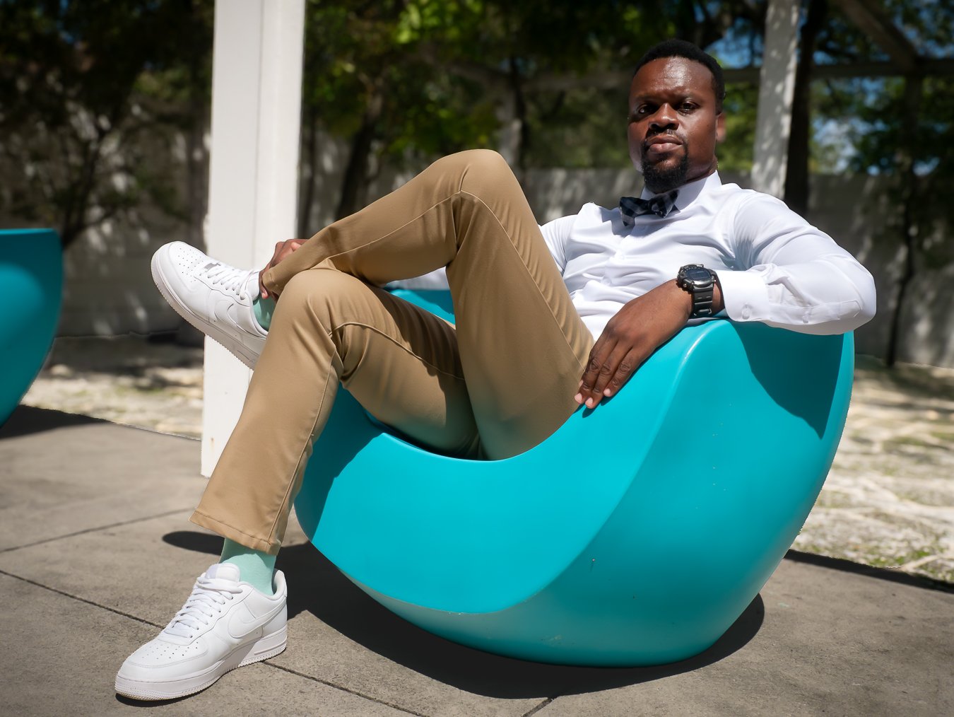Professional photographer  sitting in a modern turquoise outdoor chair on a sunny day, wearing khaki pants, a white shirt, black bow tie, white sneakers, a watch, and green socks.