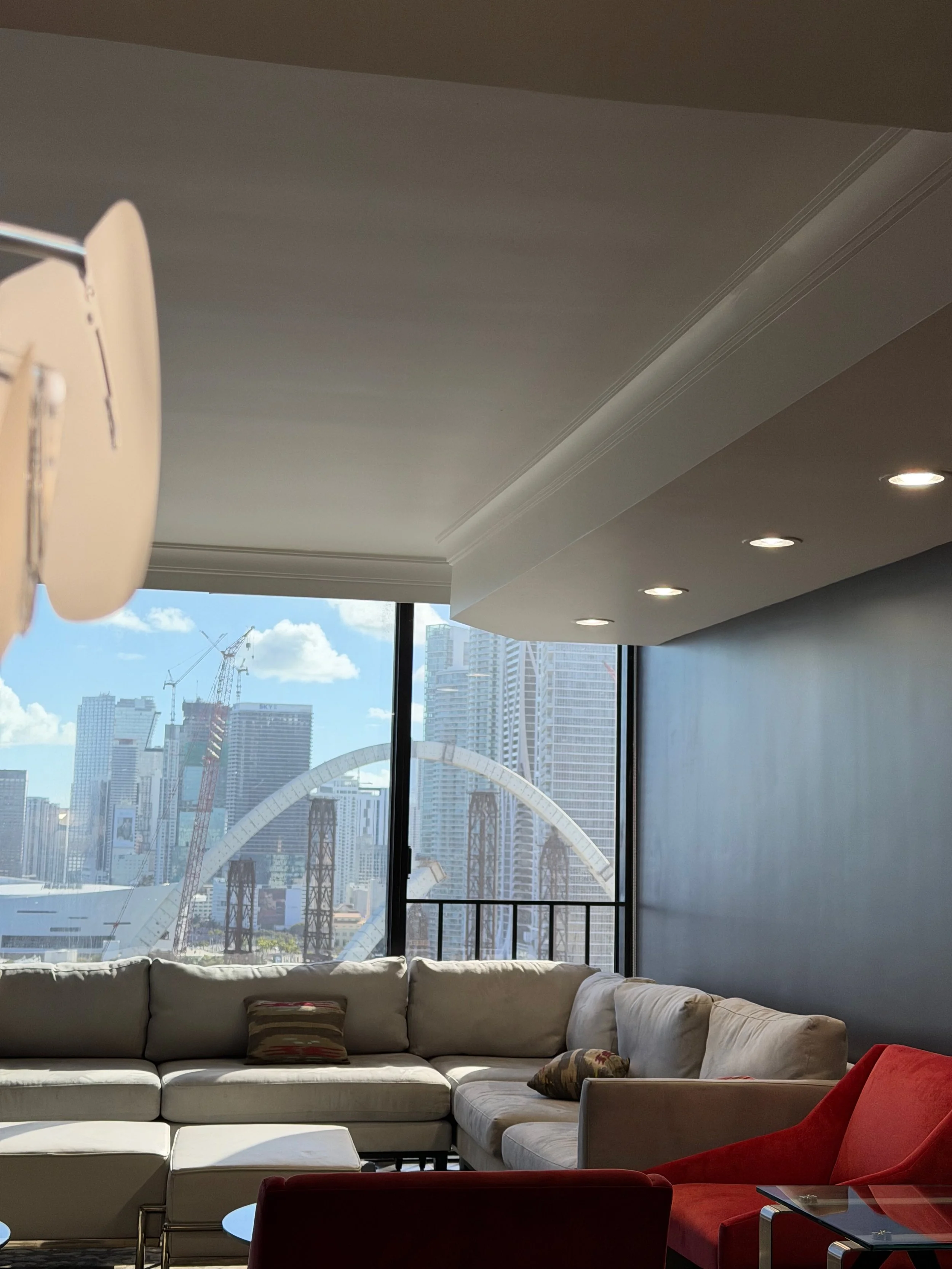 Living room with beige sectional sofa, red armchair, and view of city skyline through large window, including a Ferris wheel and construction cranes.