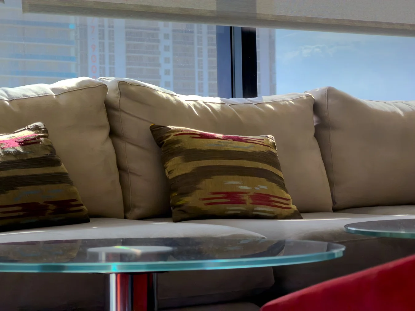 Living room sofa with decorative pillows and a glass coffee table, window with cityscape view in background.