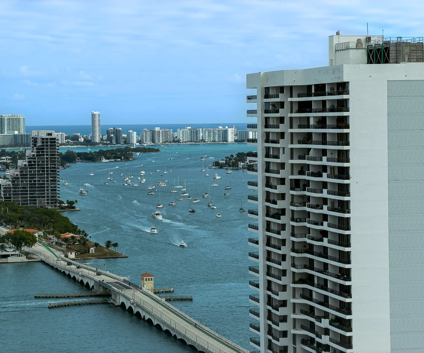 A high-rise building overlooking a waterway with numerous boats, a bridge, and a city skyline in the background.
