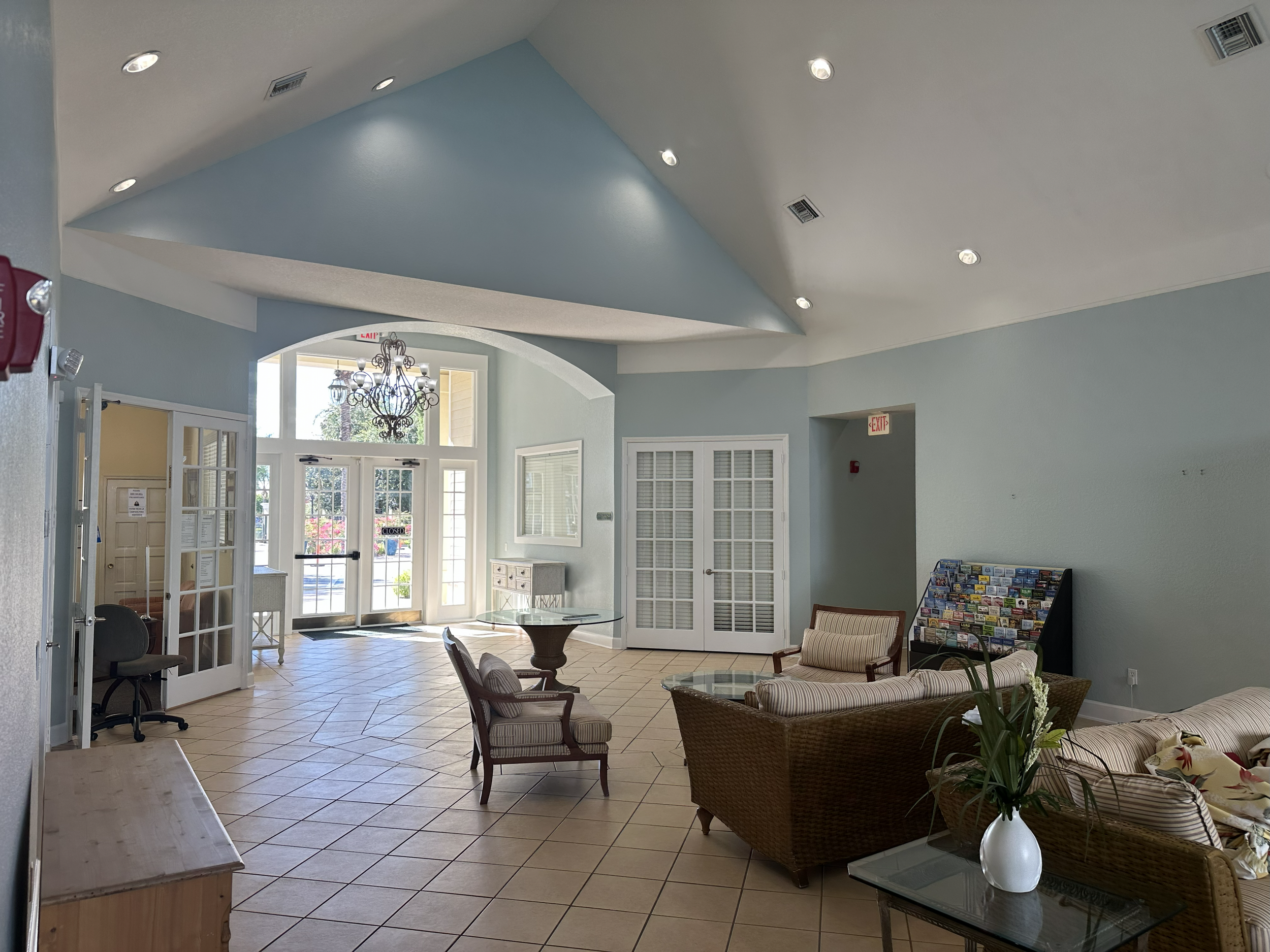 Lobby area with seating, glass tables, a chandelier, a brochure stand, and front doors with large windows.