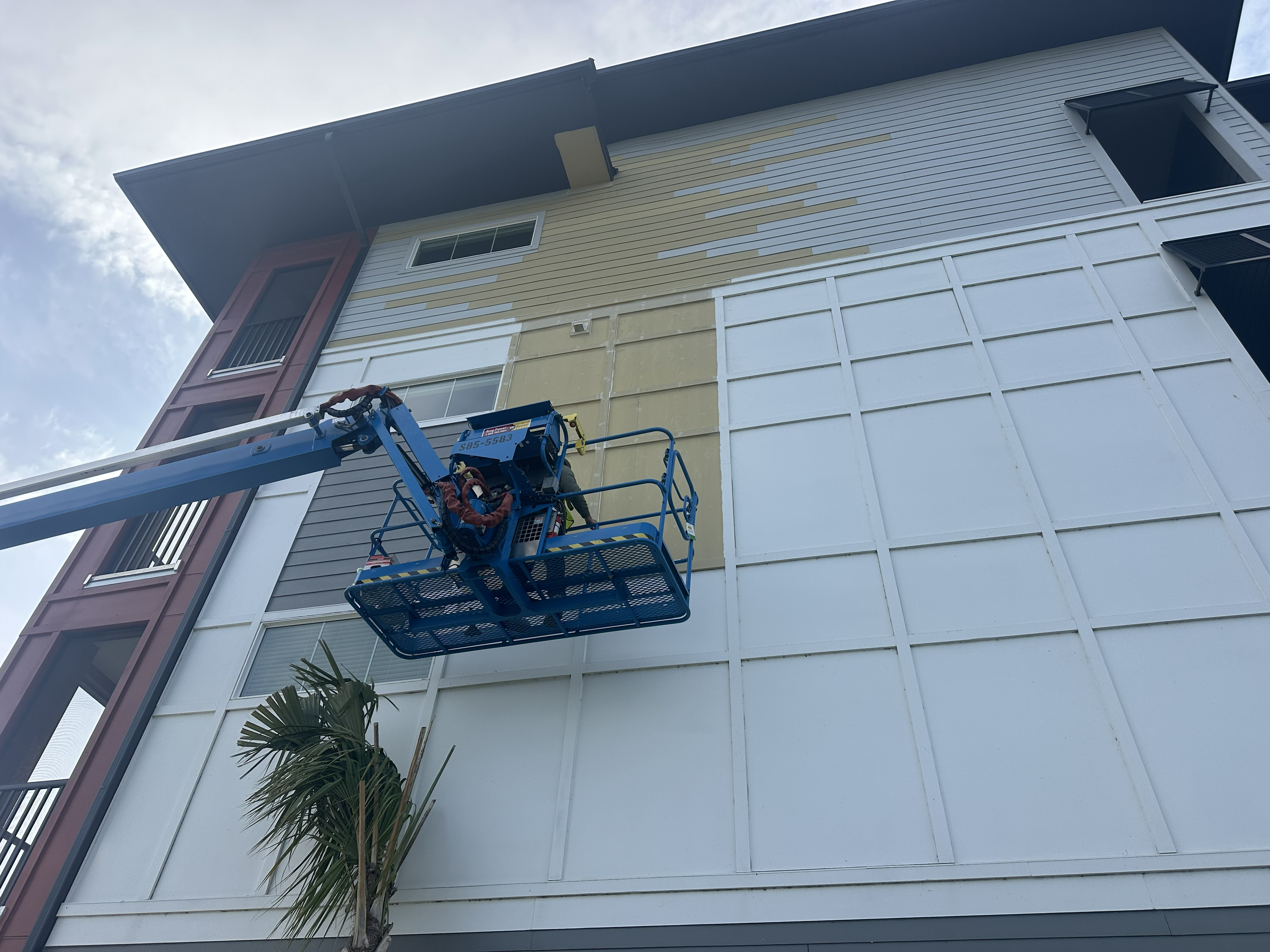 Worker in a lift painting the exterior of a multi-story building with beige and white siding.