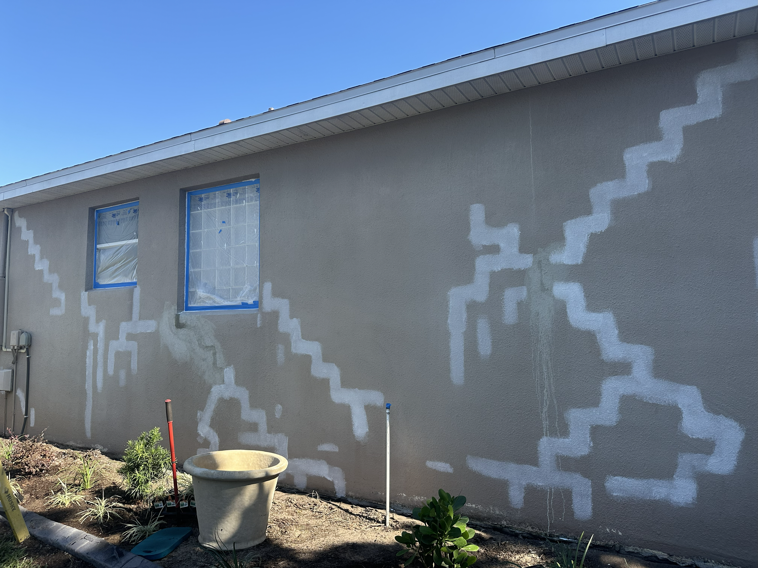 The exterior of a house under renovation with patches of primer and paint, two windows covered with plastic, and garden plants in the foreground.