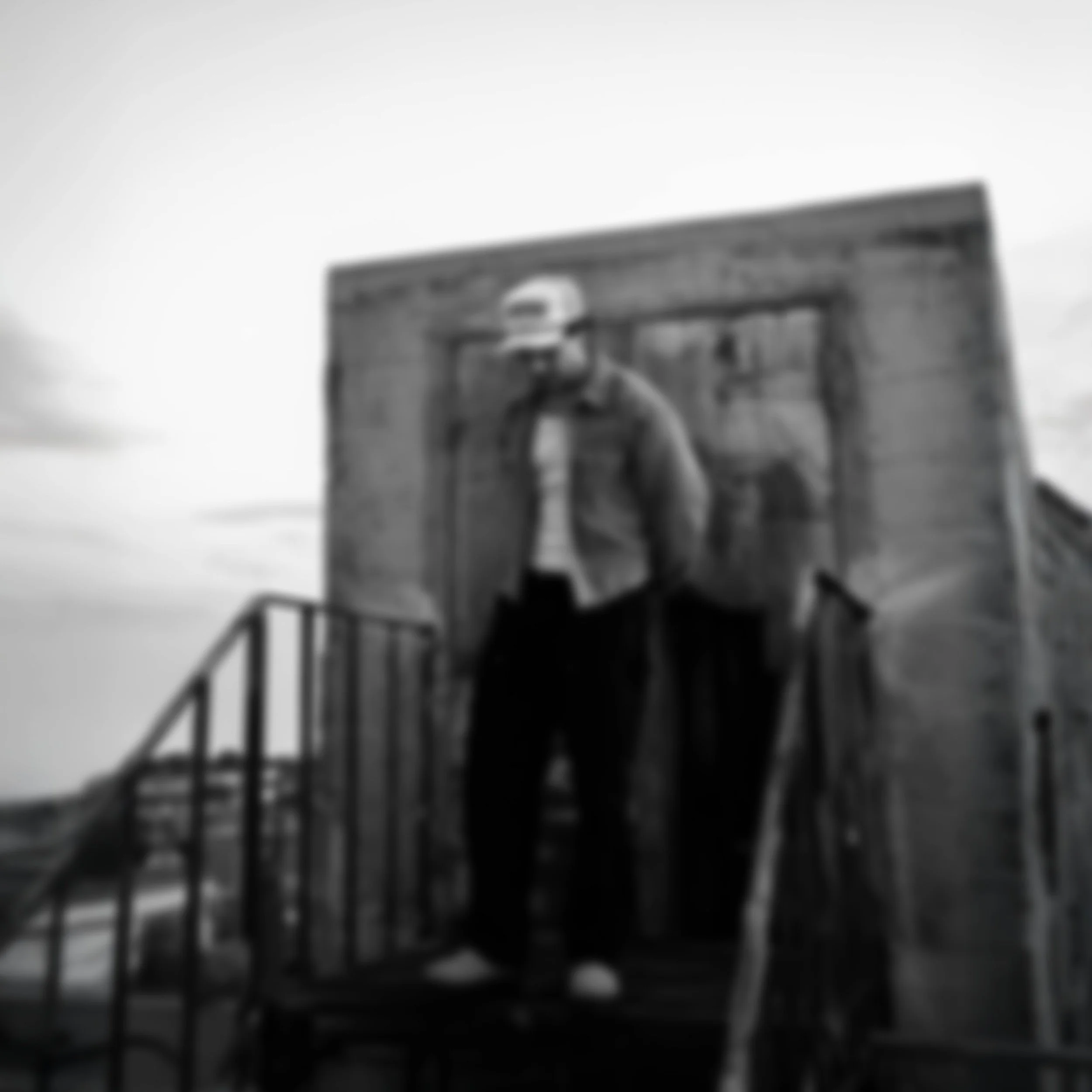 Black and white photo of a man wearing a cap, jacket, and sunglasses, standing on a staircase outside a building with a wooden door.