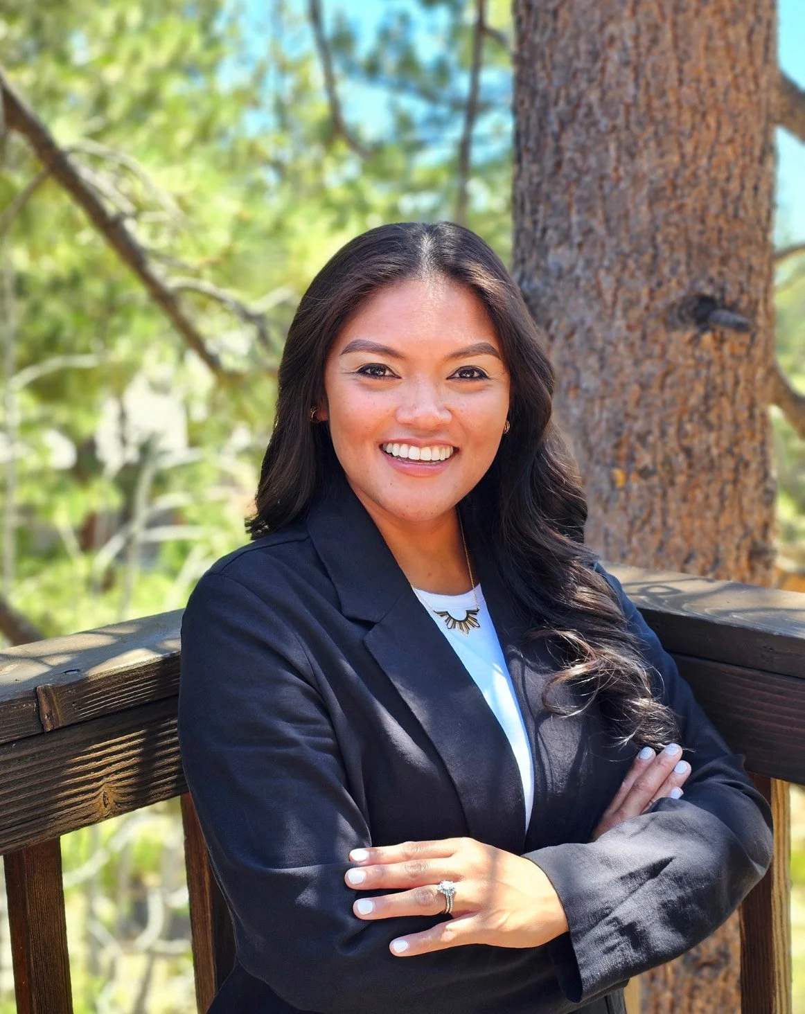 A woman with long dark hair and a white top, wearing a dark blazer, stands outdoors in front of a large tree with green leaves. She is smiling with arms crossed and is wearing a ring and a necklace. The background shows a sunny day with a partly blue sky.
