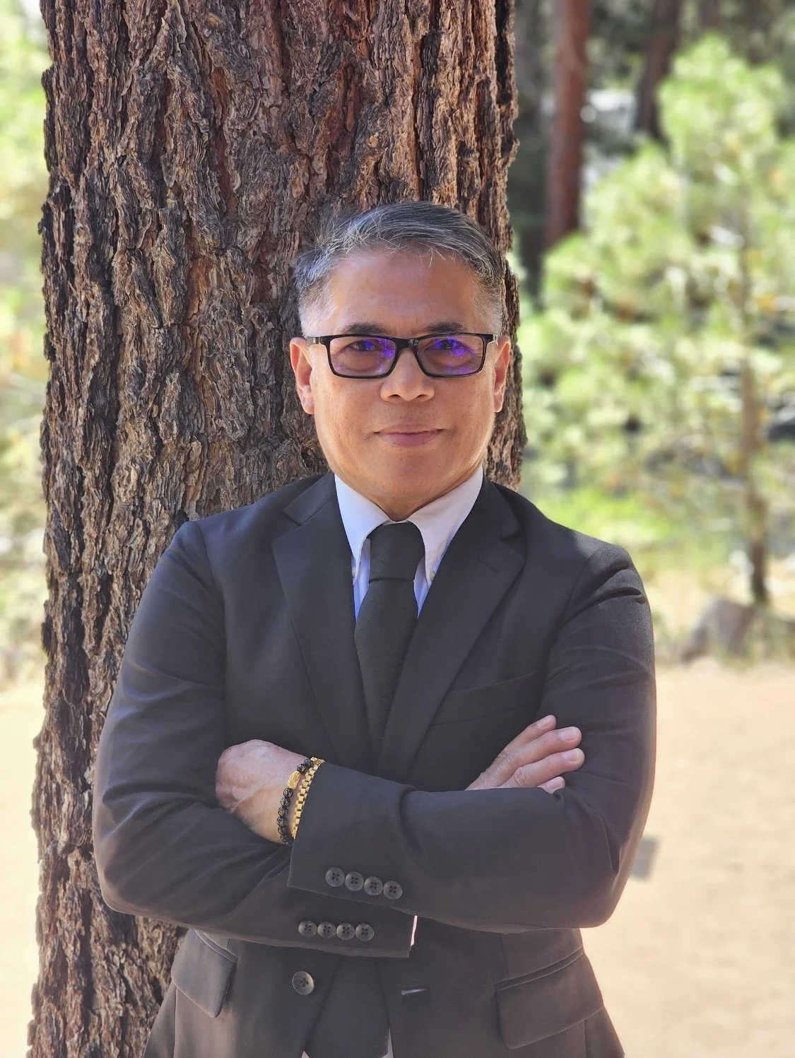 A man in a black suit and white shirt with a black tie, wearing glasses, stands with arms crossed in front of a large tree in a wooded area during daytime.