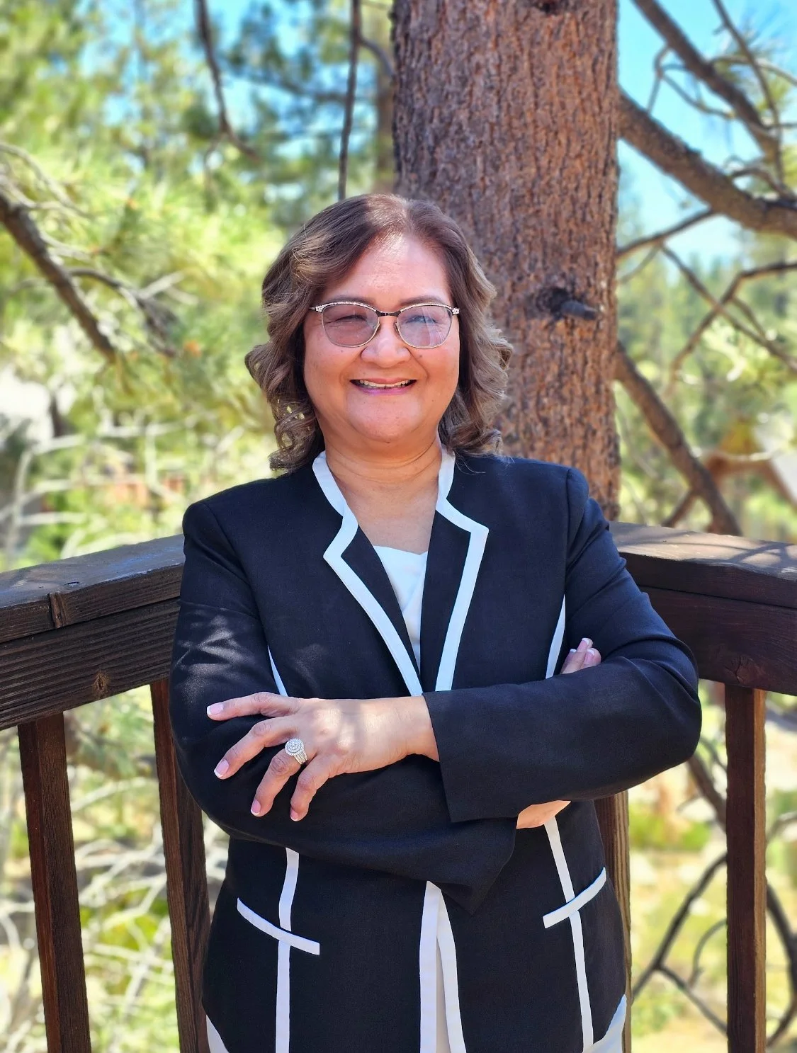 A smiling woman with glasses, shoulder-length wavy hair, wearing a black blazer with light-colored trim, standing outdoors with a wooden railing and a tree in the background.