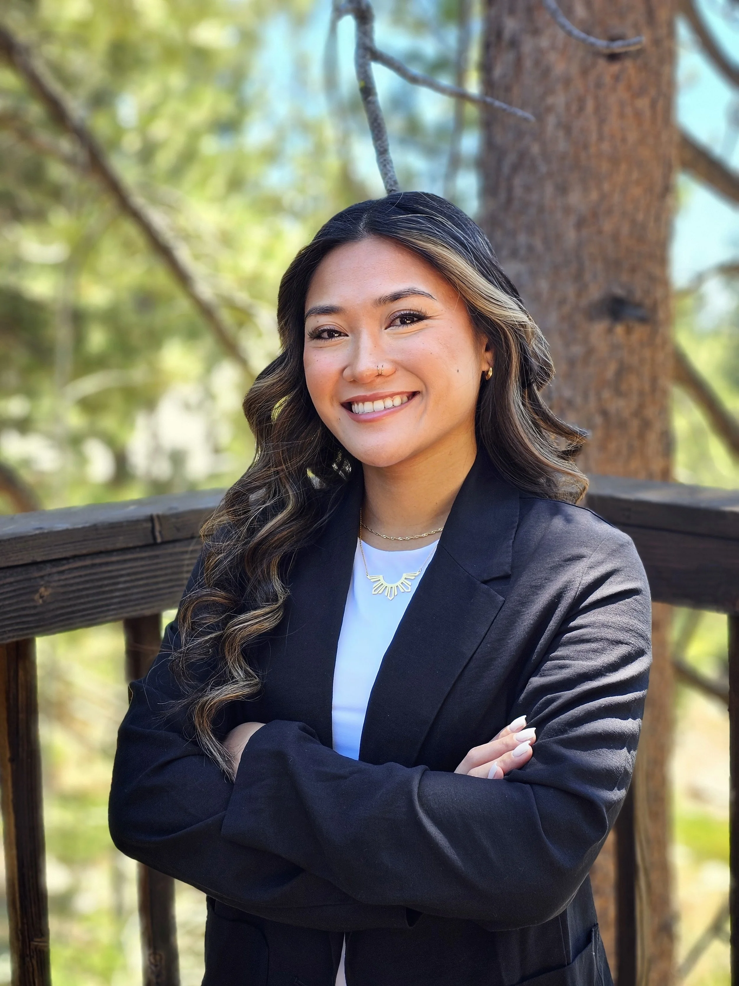A young woman with wavy dark hair and light highlights, smiling with crossed arms, wearing a black blazer, white top, gold jewelry, standing outdoors near a wooden railing with a large tree and green foliage in the background.