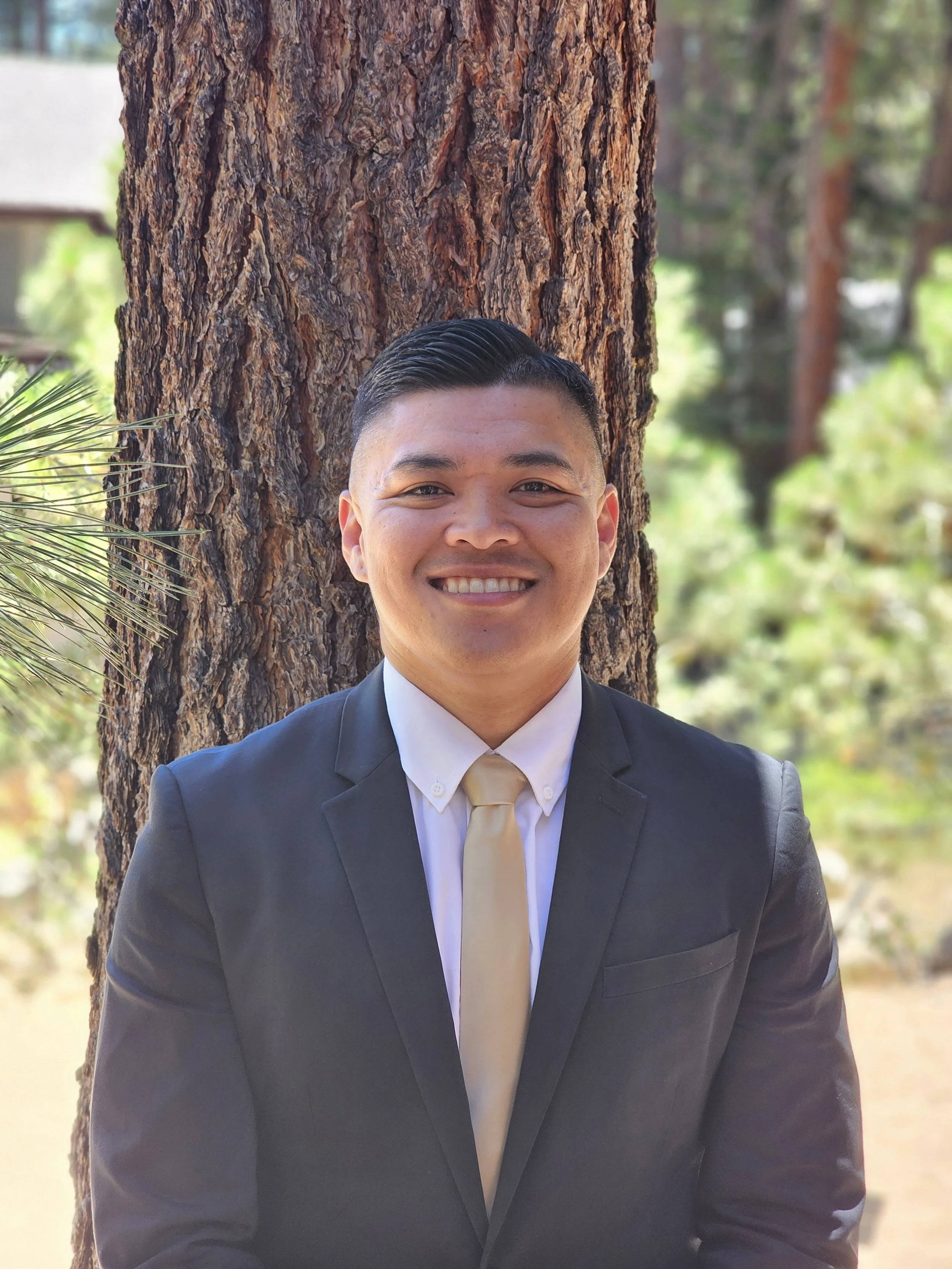 A young man in a black suit and beige tie smiling, standing in front of a large tree trunk with green foliage in the background.