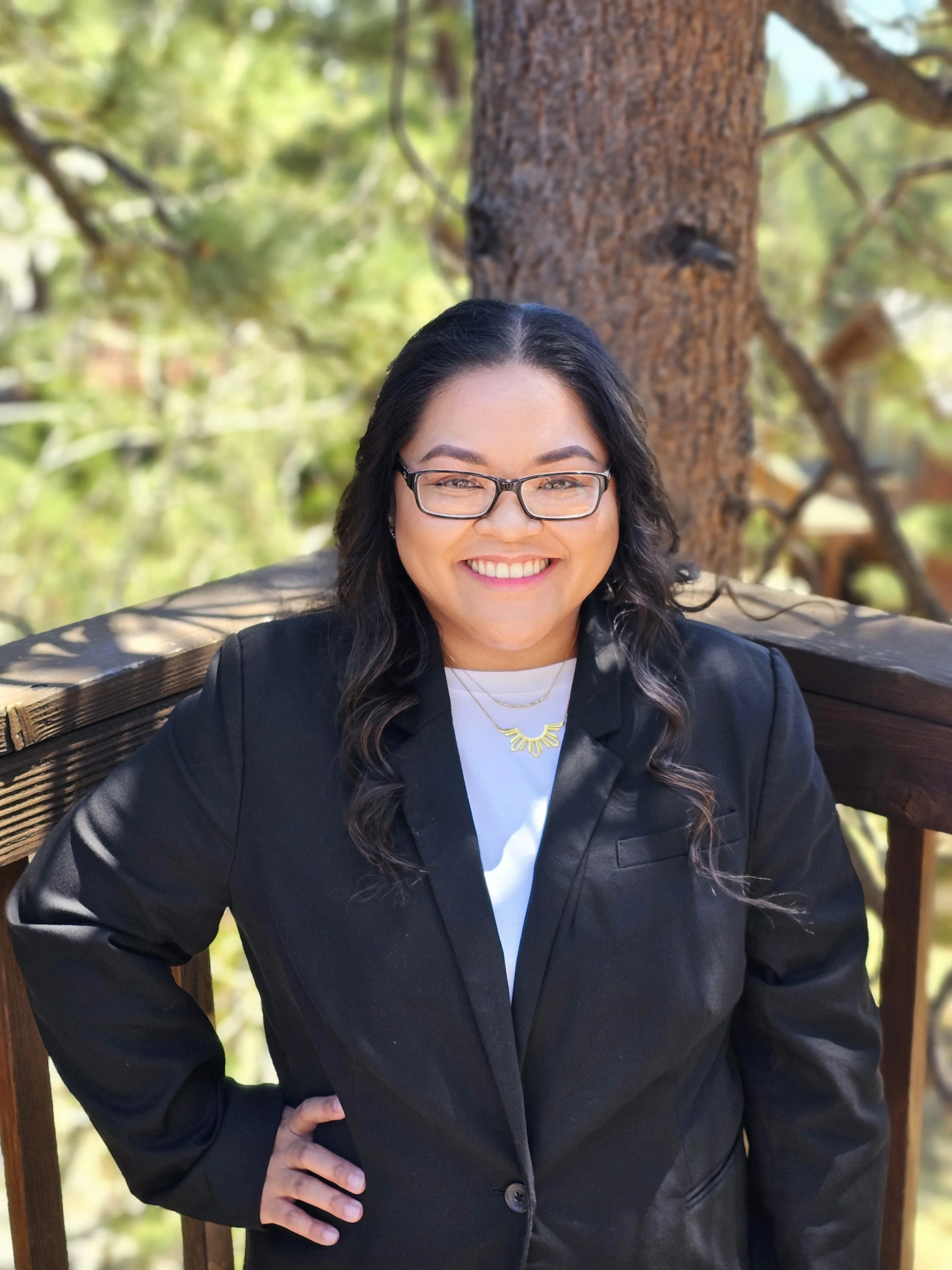 A woman with glasses, wearing a black blazer over a white shirt, standing outdoors near a tree, smiling at the camera.