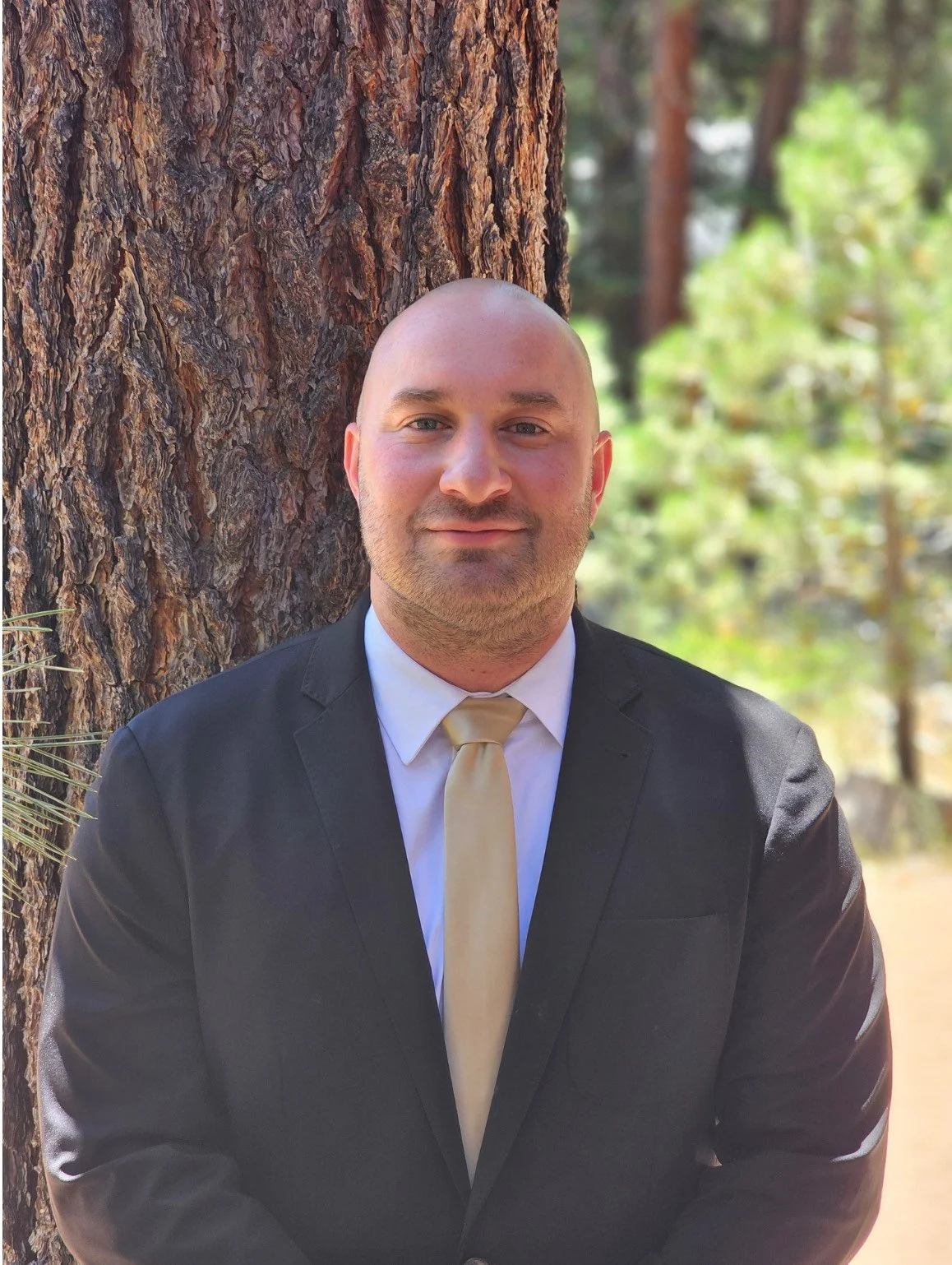 A man in a suit and tie standing outdoors next to a tree with a forest background.