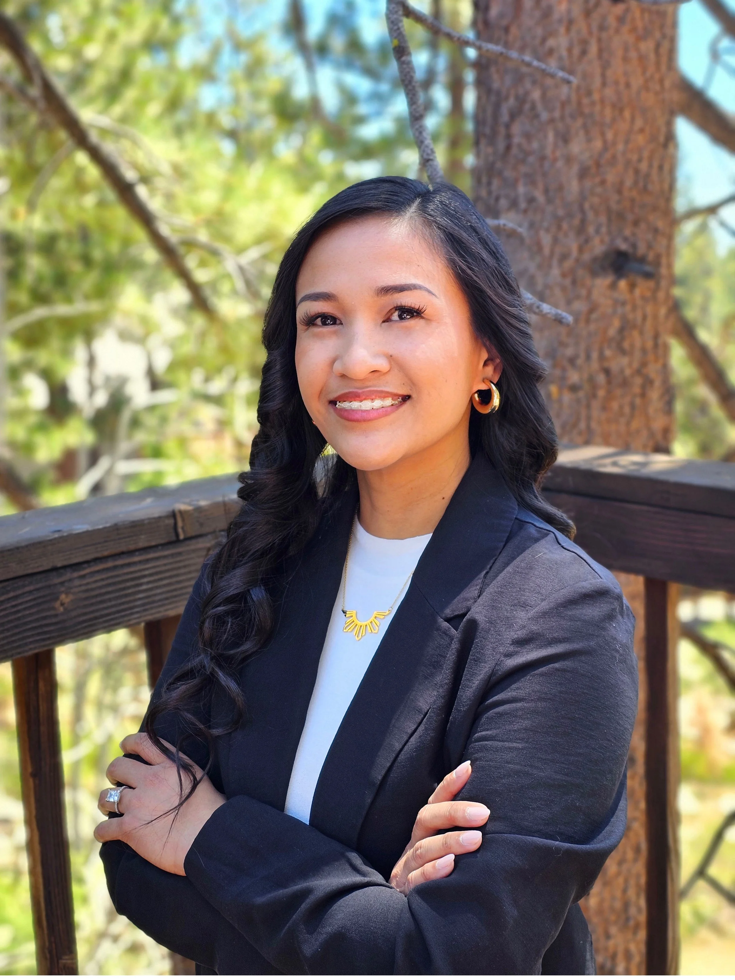 A woman with dark wavy hair, wearing a black blazer, white shirt, and gold jewelry, standing outdoors on a wooden deck with trees in the background.