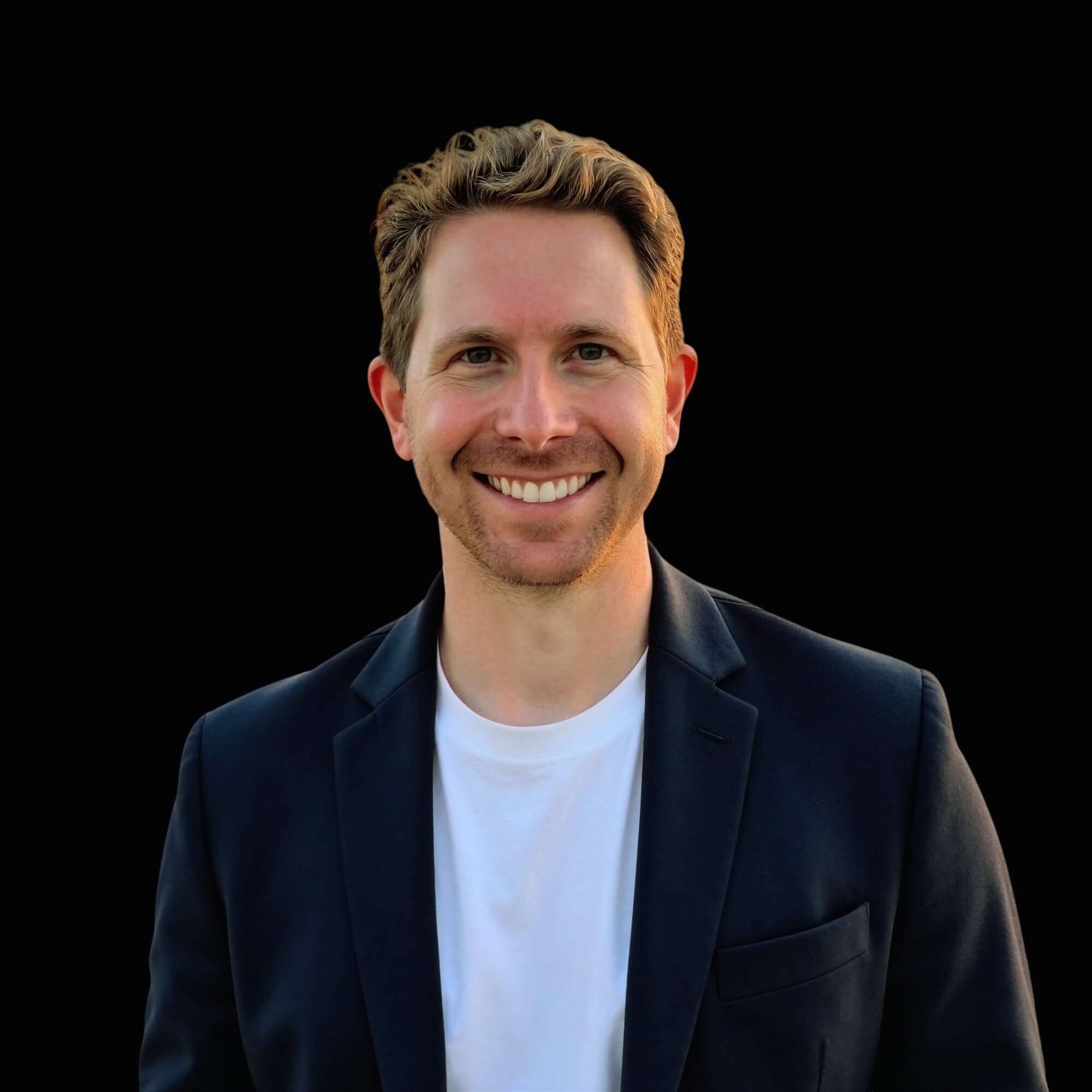 A smiling man with short wavy blonde hair, wearing a black blazer over a white shirt, standing against a solid black background.