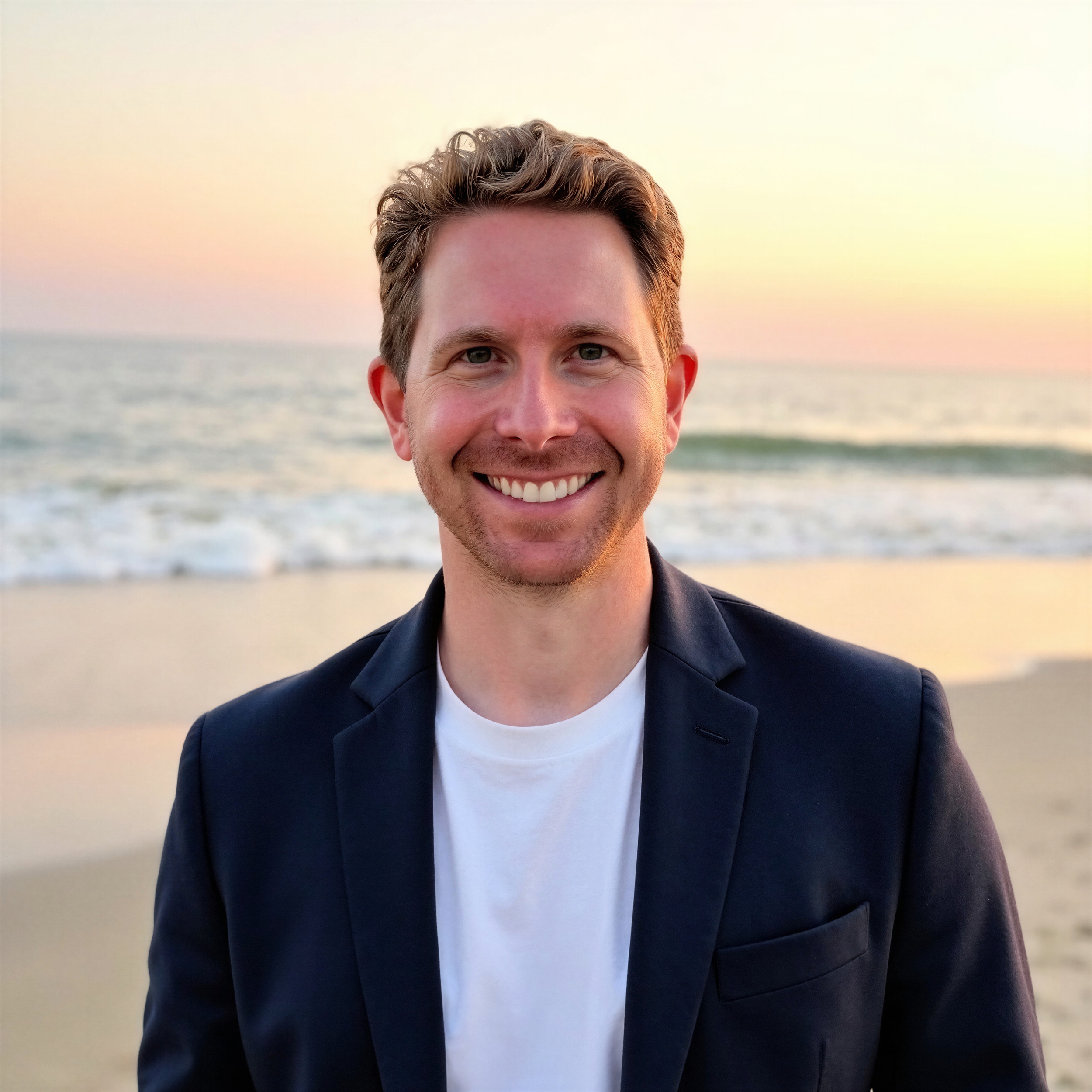 A smiling man in a dark blazer and white t-shirt stands on the beach at sunset, with the ocean and sky in the background.