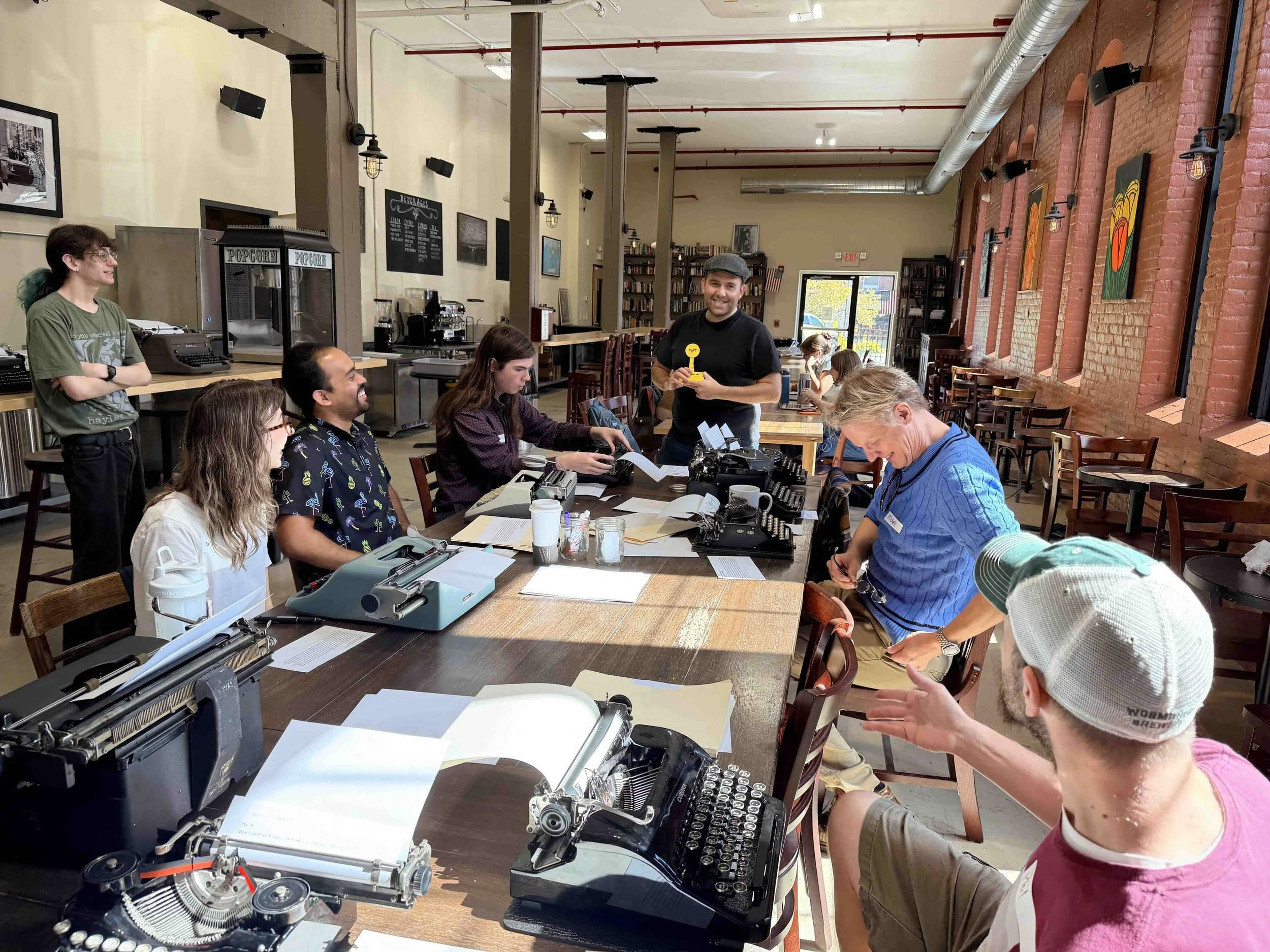 People gathered around a long table covered with vintage typewriters, paper, and writing supplies during a Tappa Tappa meetup, with one attendee standing and holding the yellow Bent Key speed challenge trophy.