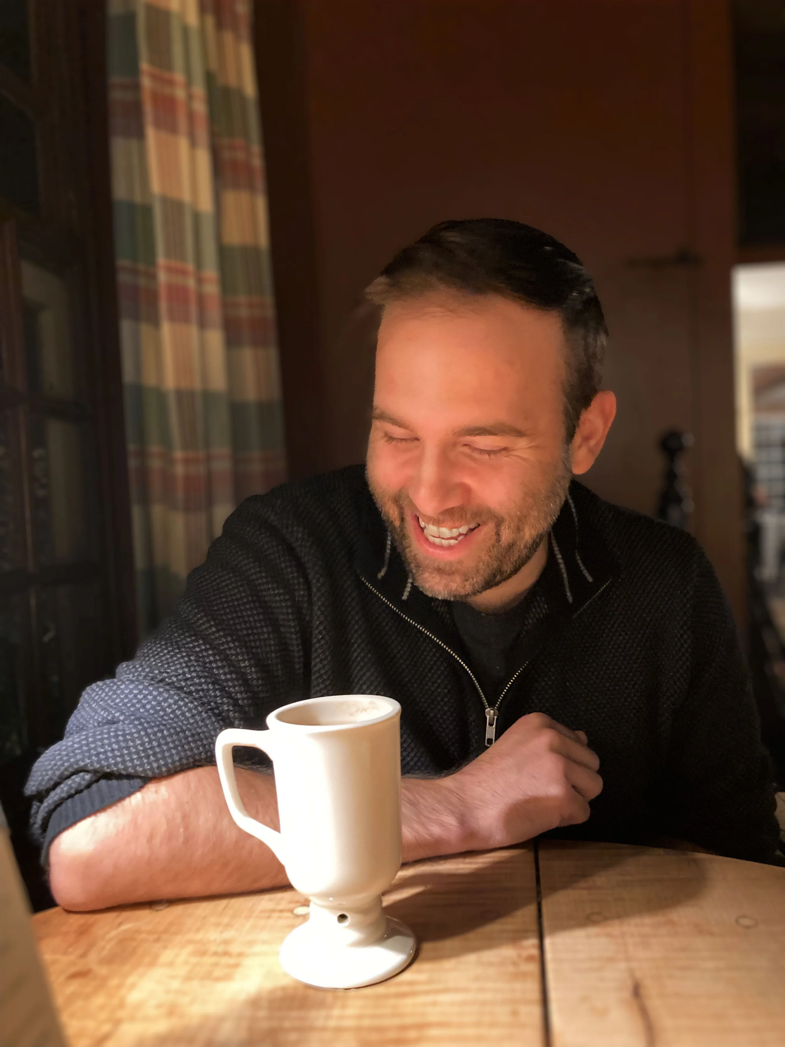A man with dark hair and a beard is smiling and looking down at a white mug of coffee or tea on a wooden table. He is wearing a black zip-up sweater and sitting in a warmly lit room with dark wood paneling and patterned curtains in the background.